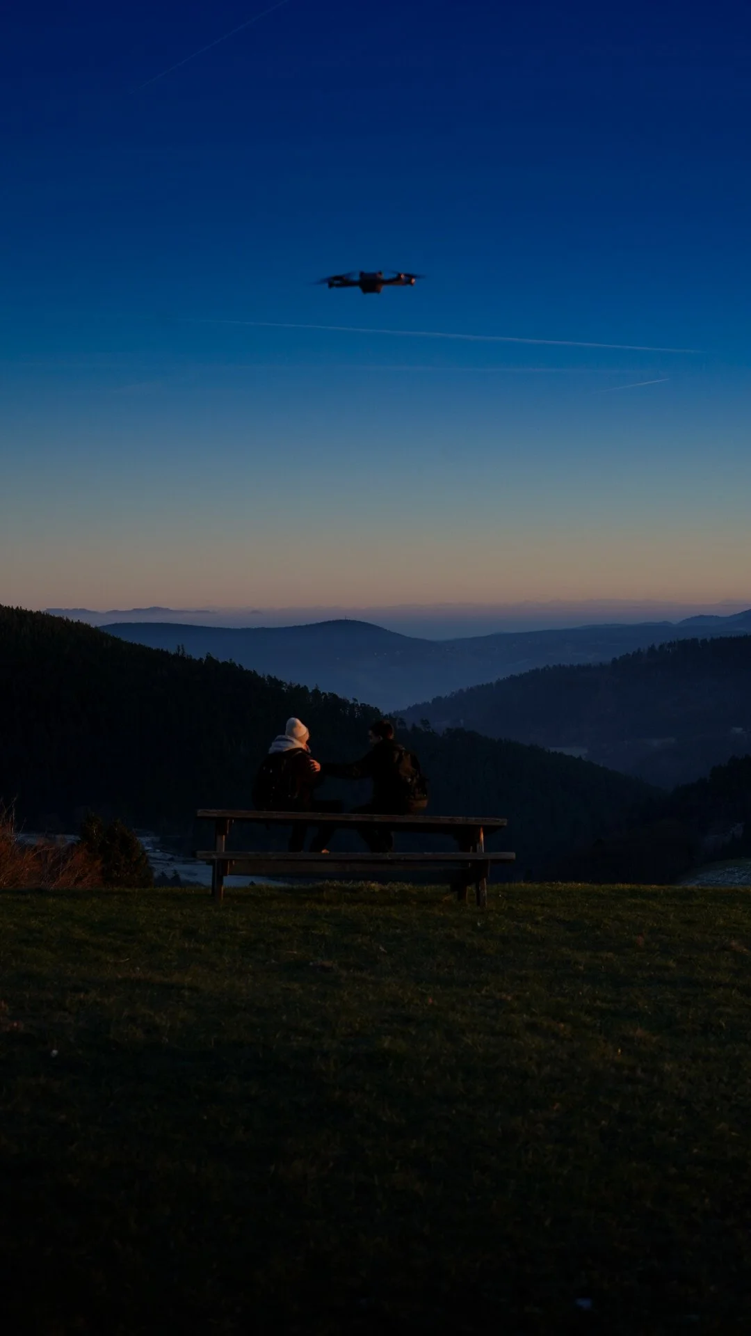 Deux personnes assises sur un banc regardant un drone dans le ciel au coucher du soleil, avec des montagnes en arrière-plan.