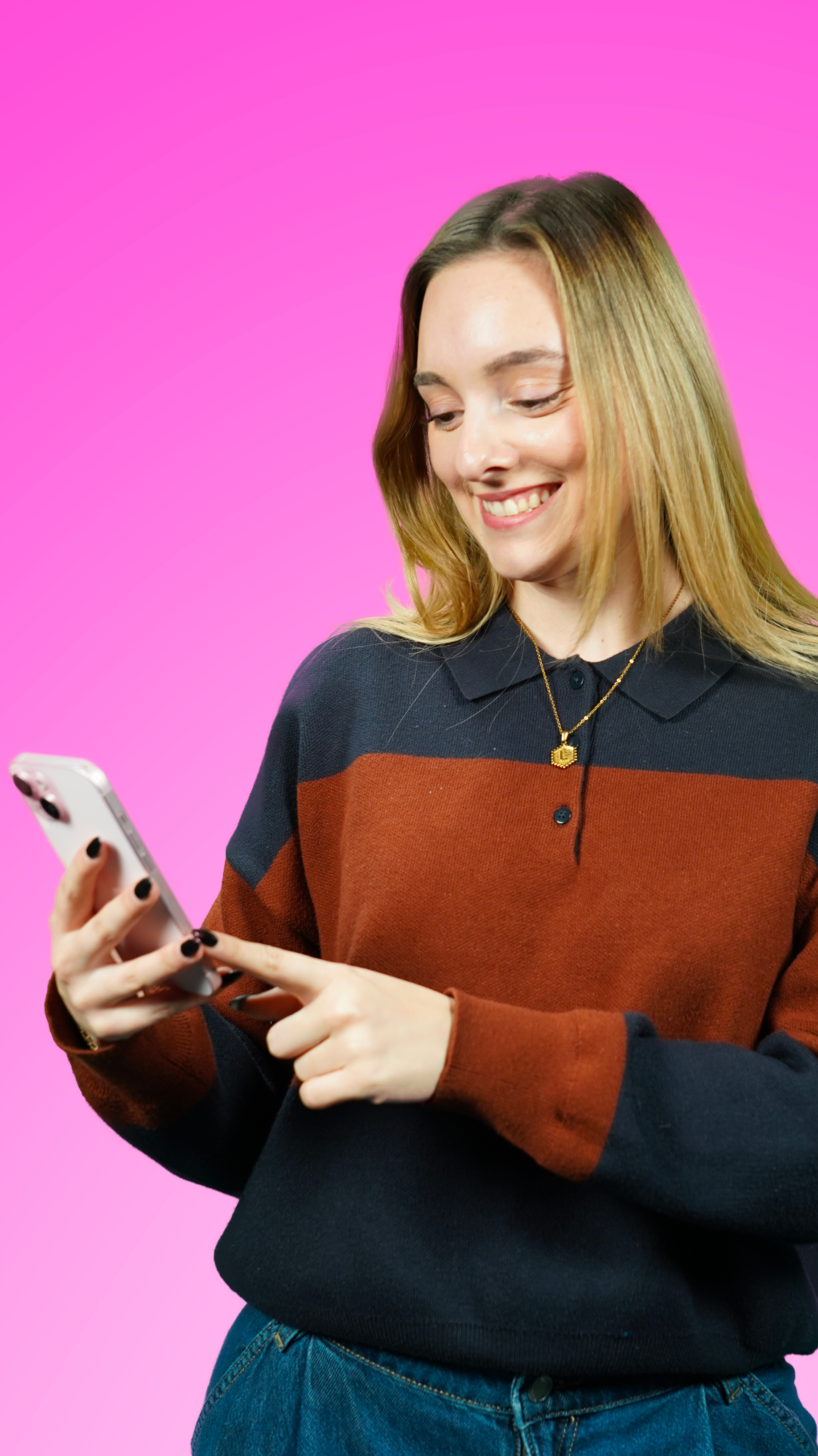 Jeune femme souriante regardant son téléphone portable, portant un pull à rayures et un collier doré, sur fond rose