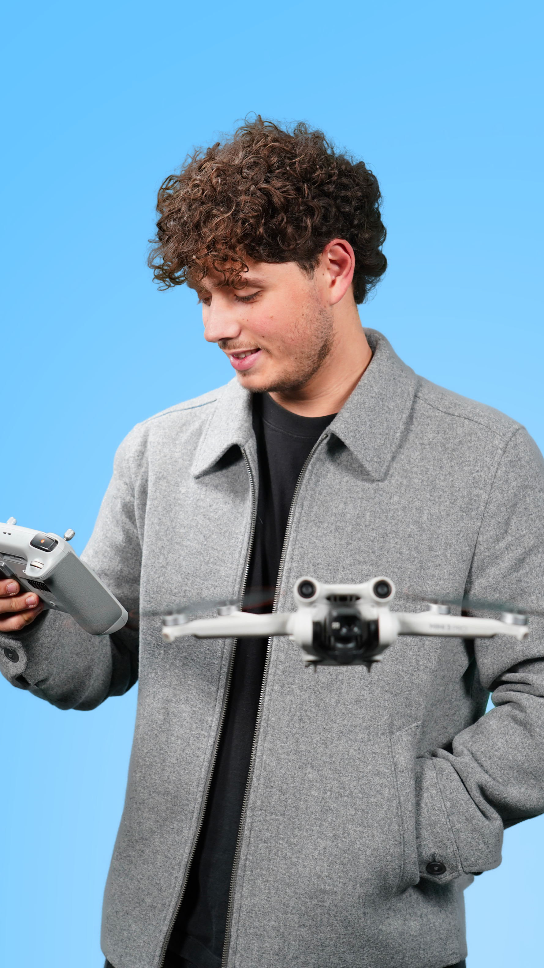 Jeune homme avec cheveux bouclés, portant un manteau gris, regarde un drone blanc et une télécommande, contre un fond bleu.