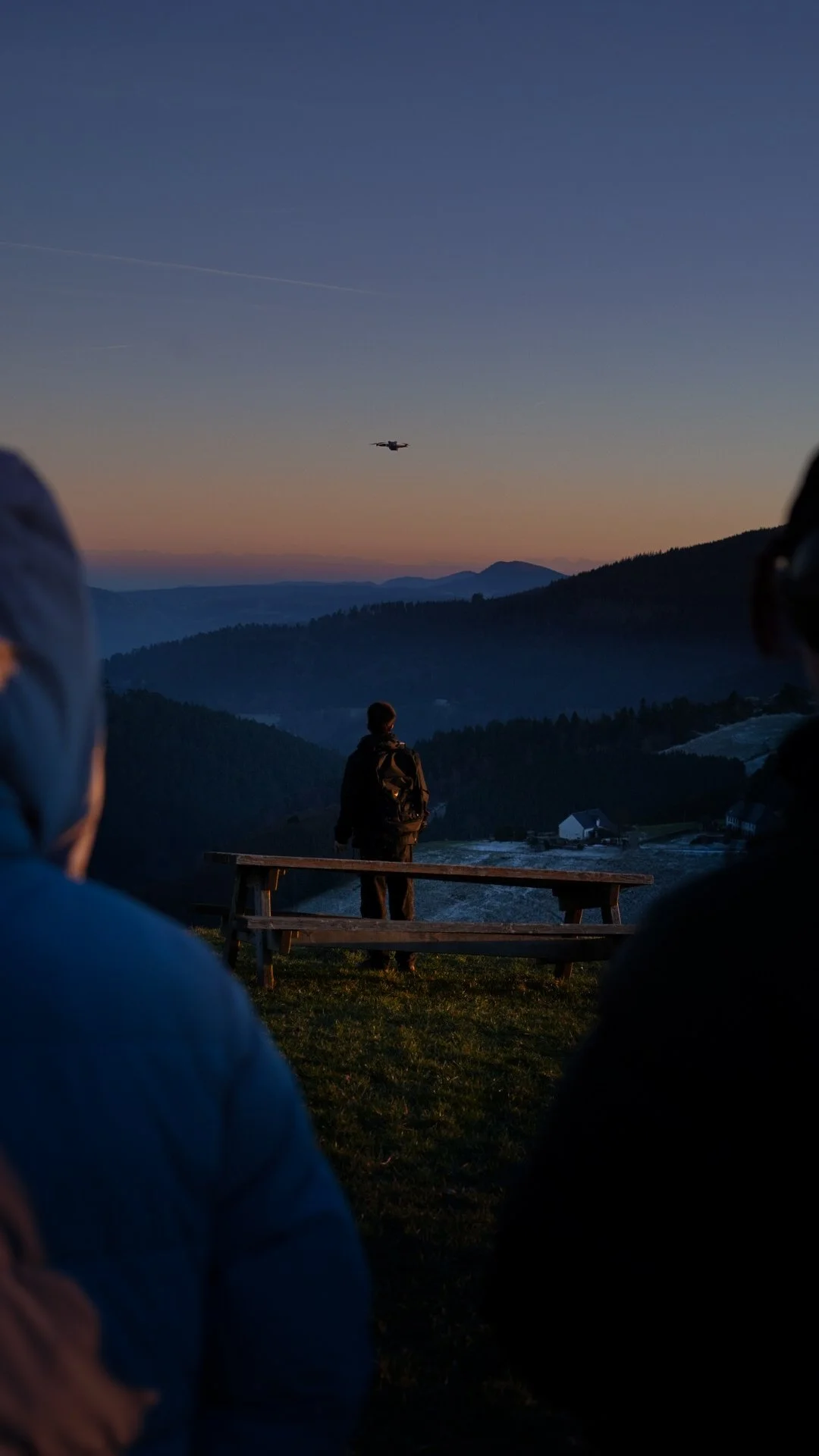Personne debout sur un banc en regardant un avion dans le ciel au coucher du soleil, entourée de montagnes et de deux personnes assises au premier plan.