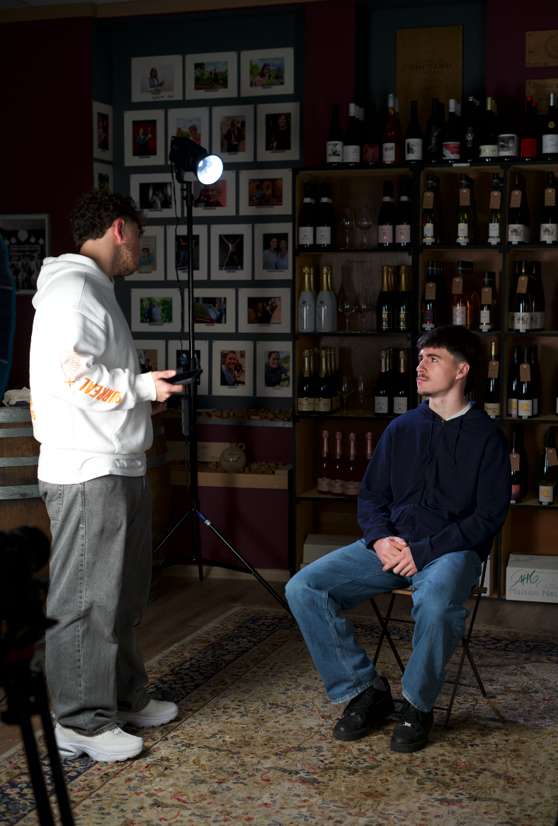 Une scène de tournage en intérieur dans un magasin de vin, avec deux jeunes hommes, l'un assis et l'autre debout. Le jeune homme debout tient un téléphone, la scène est éclairée par une lampe spécialisée.