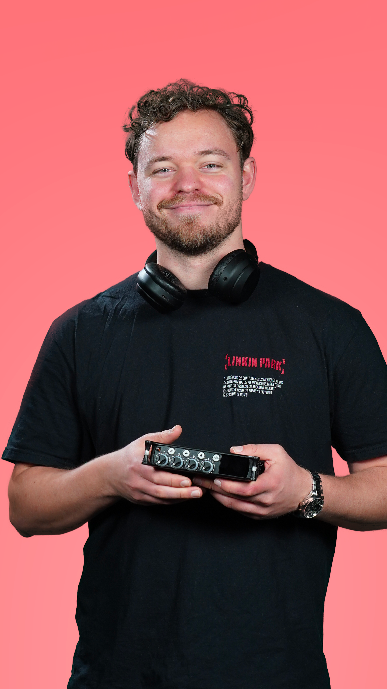 Jeune homme souriant portant des écouteurs autour du cou et tenant un petit équipement audio, portant un t-shirt noir avec le logo Pink Floyd, contre un fond rose.|