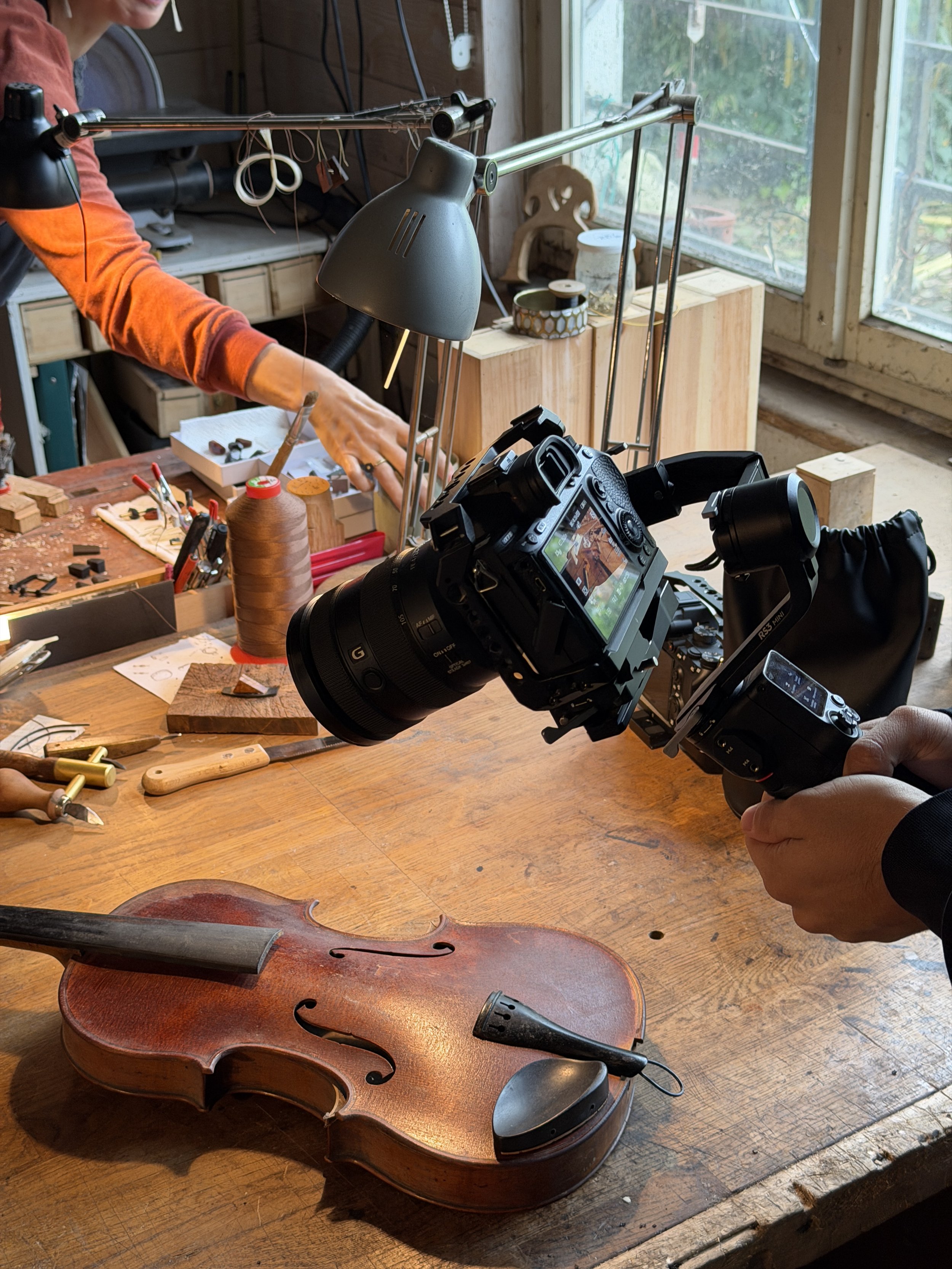 Un atelier de luthier avec un violon posé sur une table en bois, un appareil photo professionnel pointé vers l'instrument, et divers outils de travail.