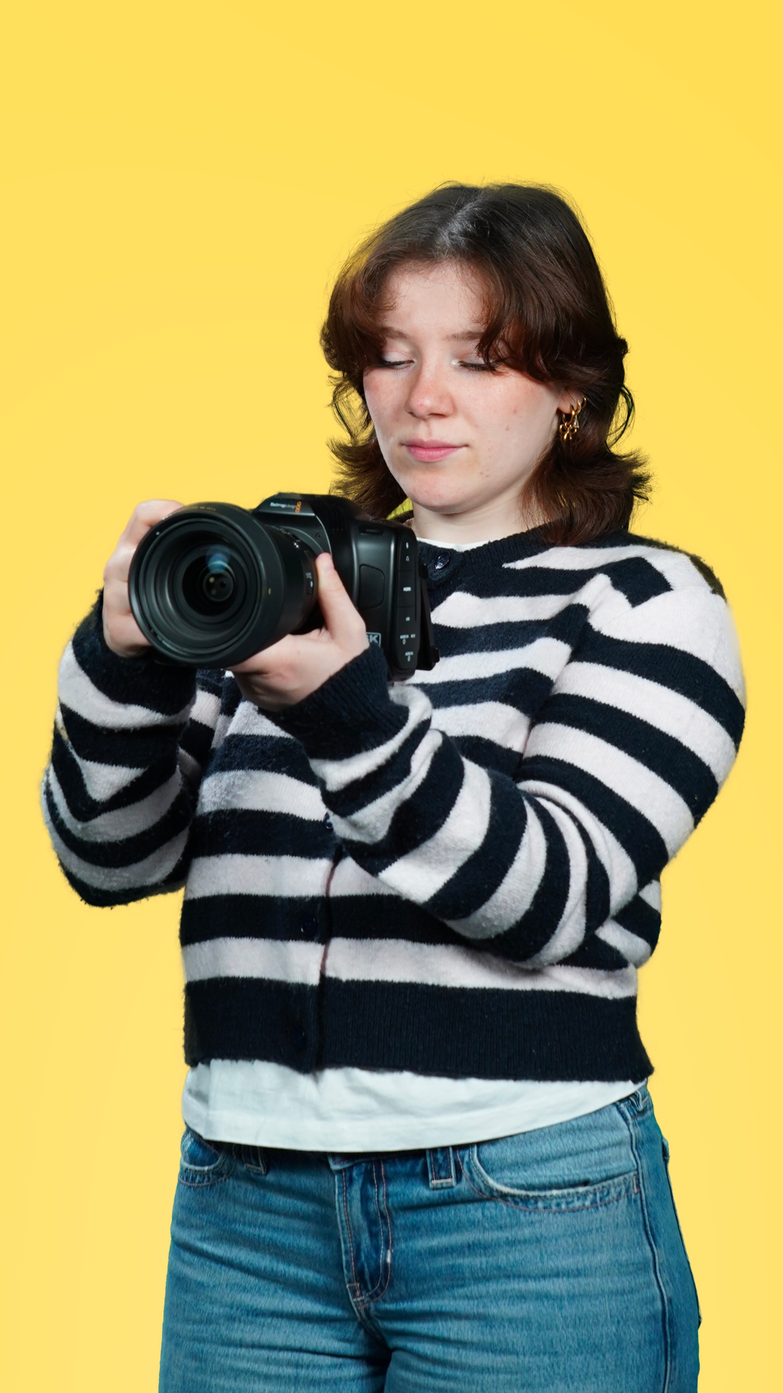 Une jeune femme avec des cheveux marron, portant un pull à rayures noires et blanches, tient un appareil photo numérique, devant un fond jaune.