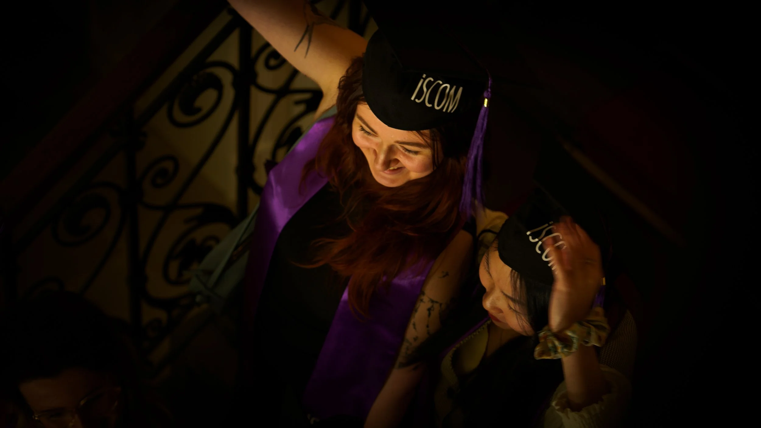 Une femme portant une coiffe de graduation avec le texte 'ISC' et une robe violette, souriante, vue de dessus dans un espace intérieur avec des escaliers en fer forgé.