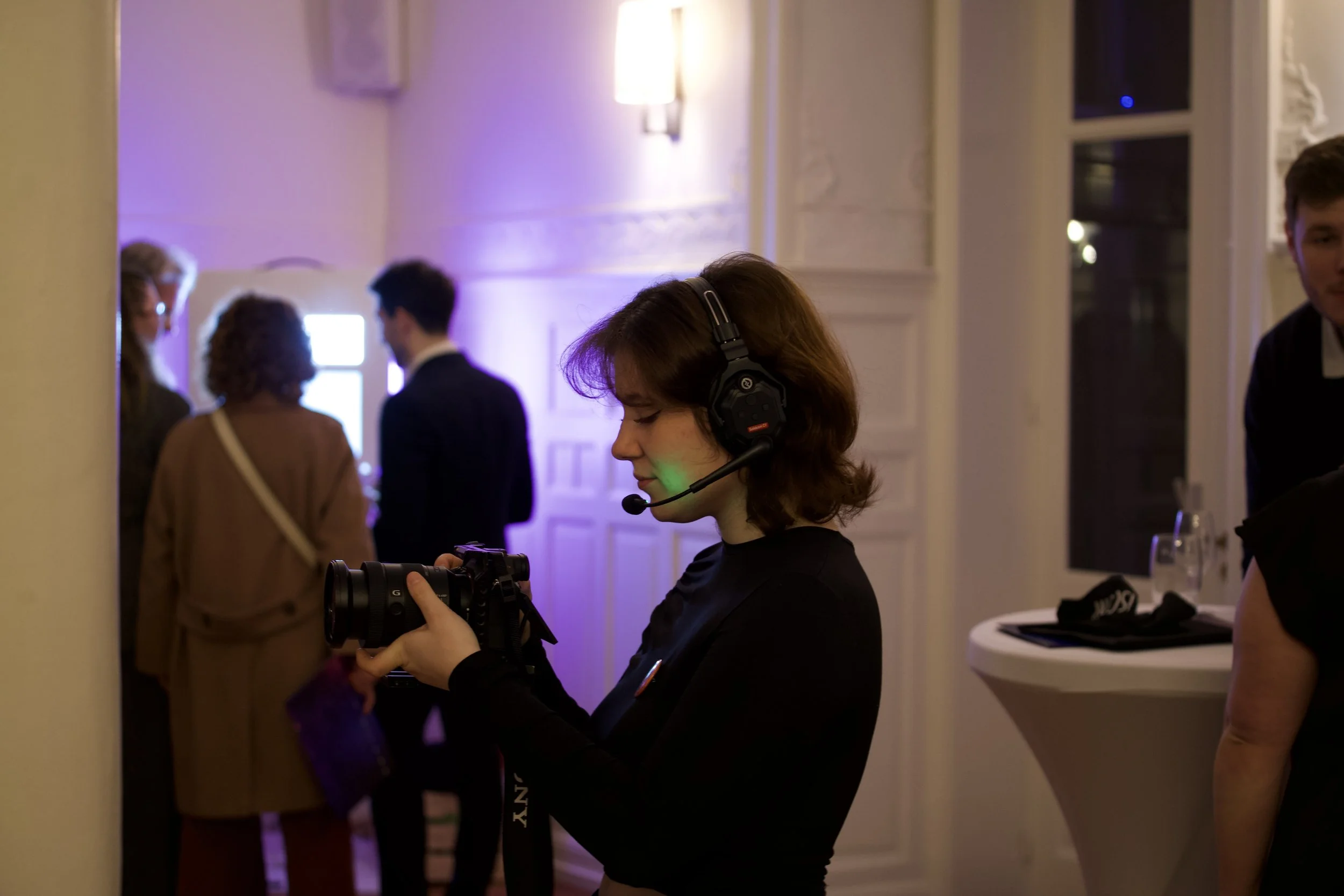 Une femme portant un casque d'écoute avec micro manipule une caméra lors d'un événement en intérieur, avec plusieurs personnes en arrière-plan