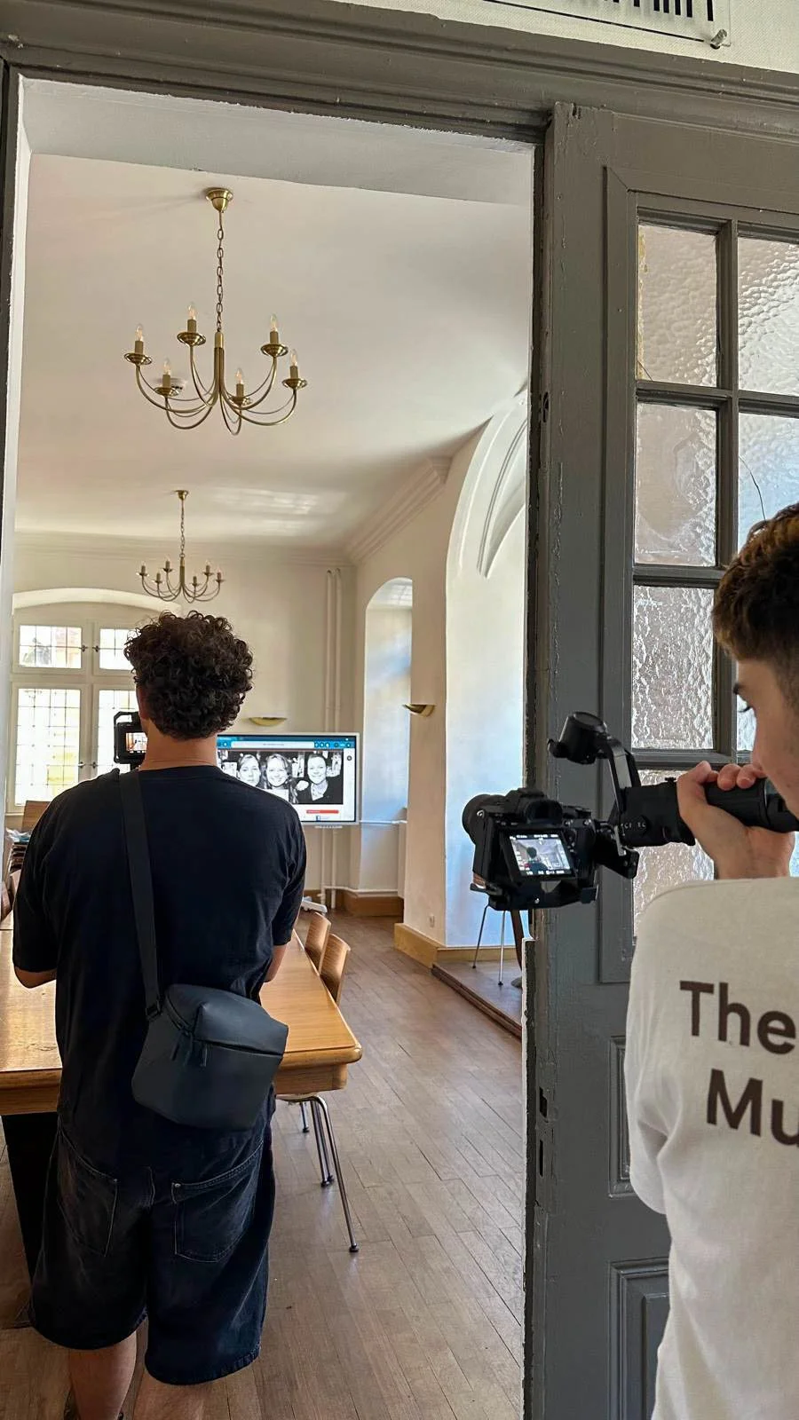 Deux jeunes hommes tournant une vidéo ou prenant une photo dans une salle avec un grand écran montrant des images de visages, dans une pièce lumineuse avec des lustres et des fenêtres.