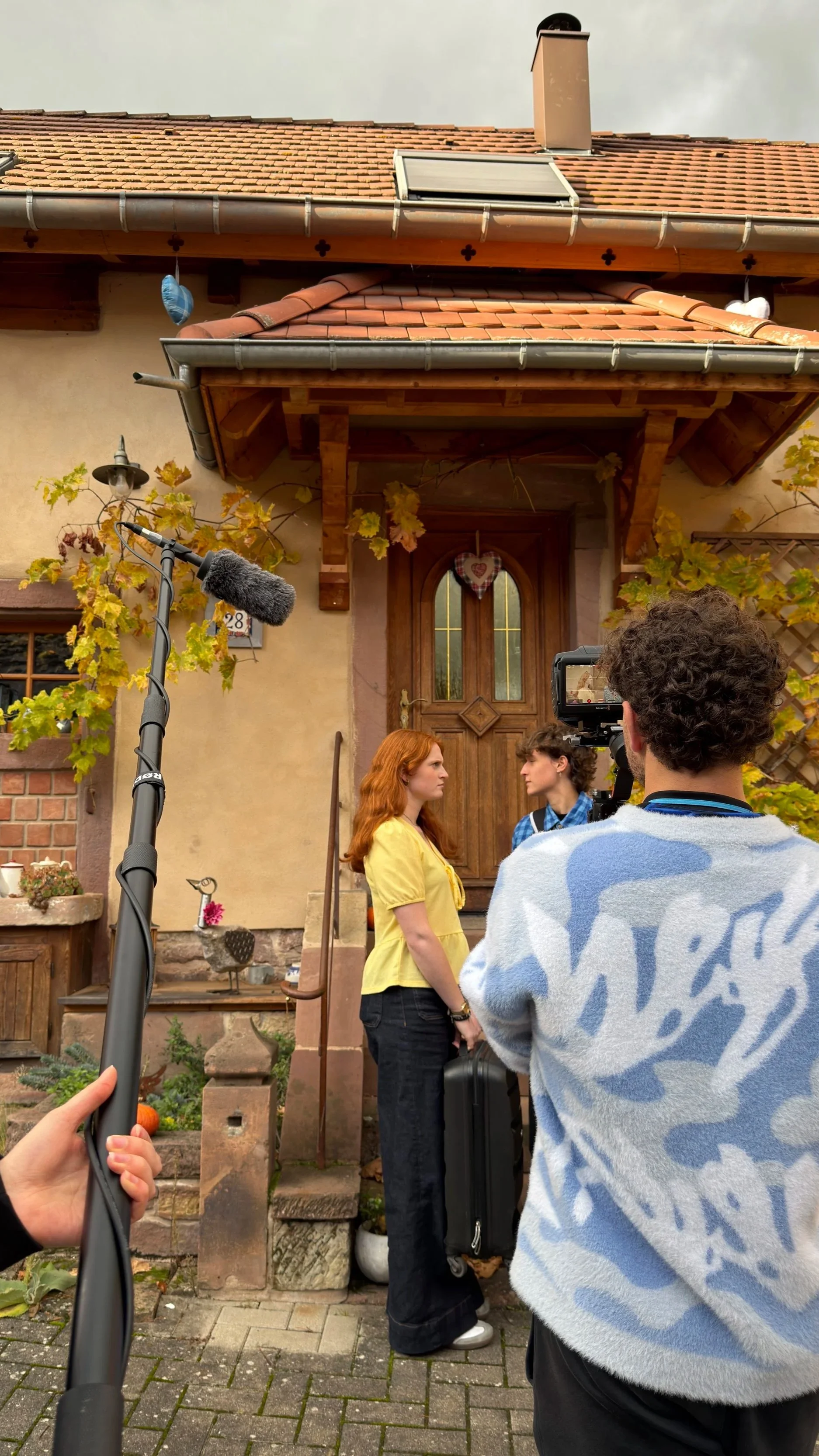 Une scène de tournage devant une maison en pierre avec deux femmes parlant, une personne derrière la caméra, un micro au premier plan, et un décor de jardin avec des plantes et des décorations.