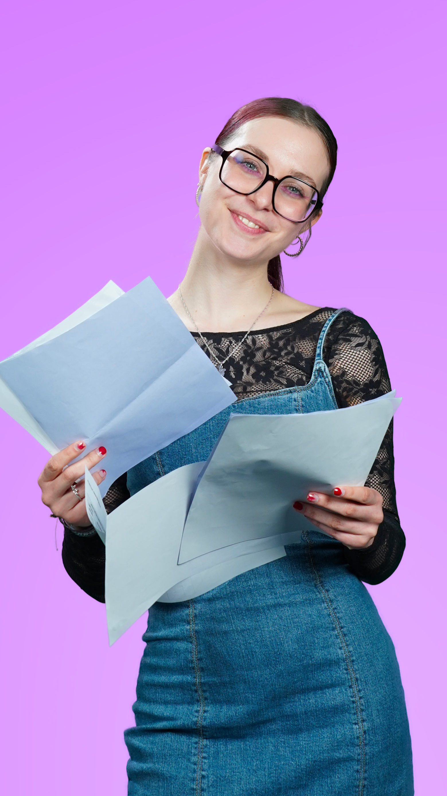 Jeune femme souriante tenant plusieurs papiers, portant des lunettes, un haut en dentelle noir et une salopette en jean, devant un fond violet.