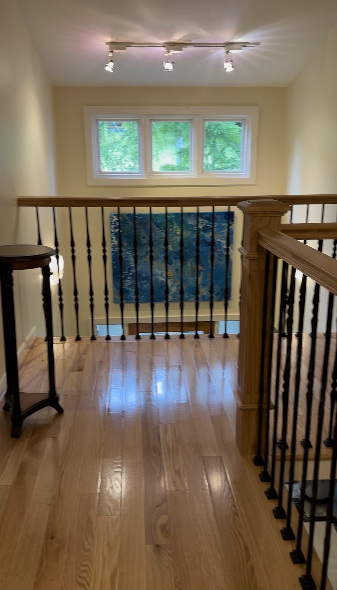 Inside view of a stair landing with three small windows, a blue abstract painting, a wooden handrail, a chandelier with three lights, and a small side table.