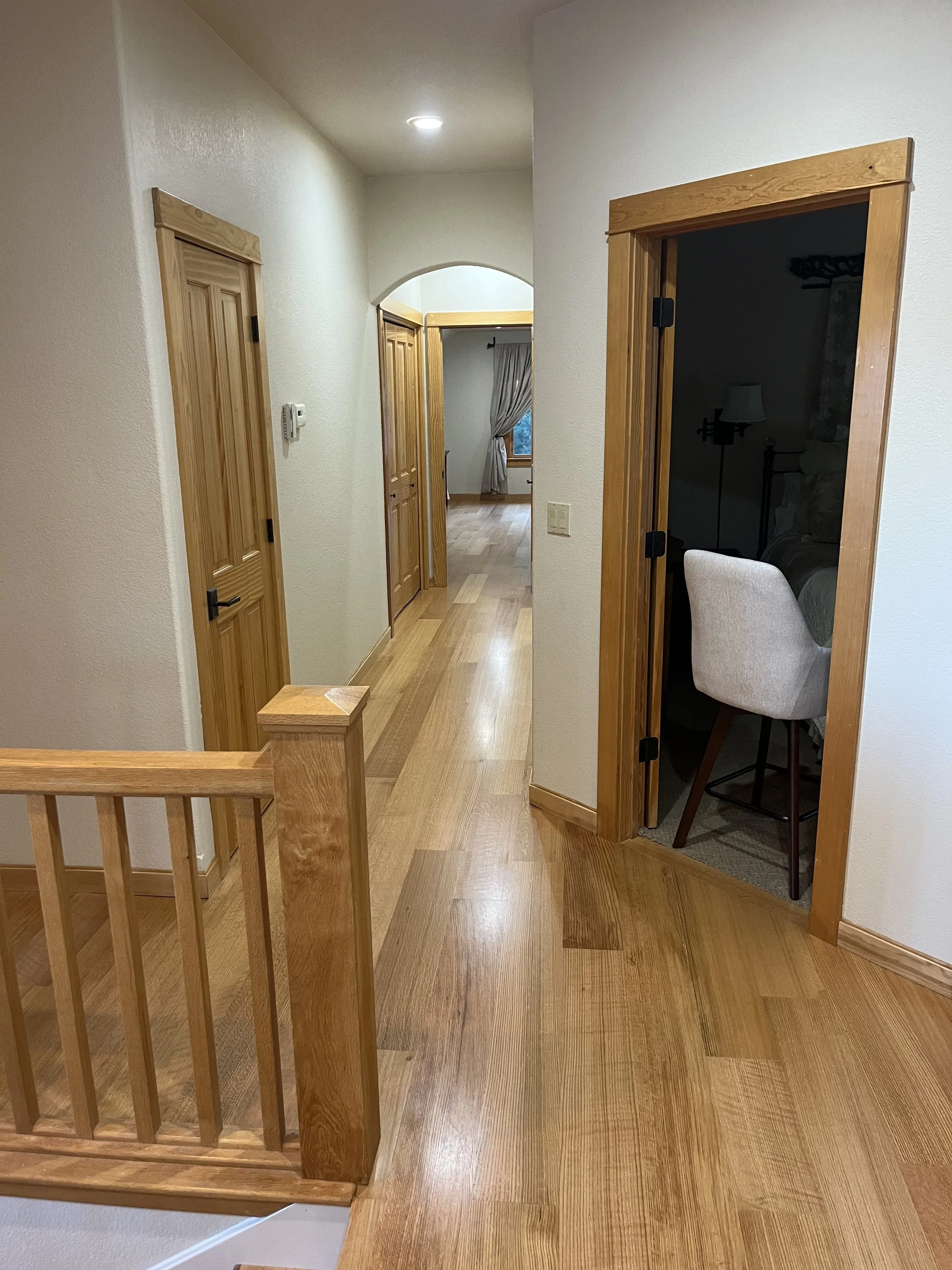 A hallway with wooden floors and doors, a window with curtains, and a doorway leading into a dark room with a lamp and chair.