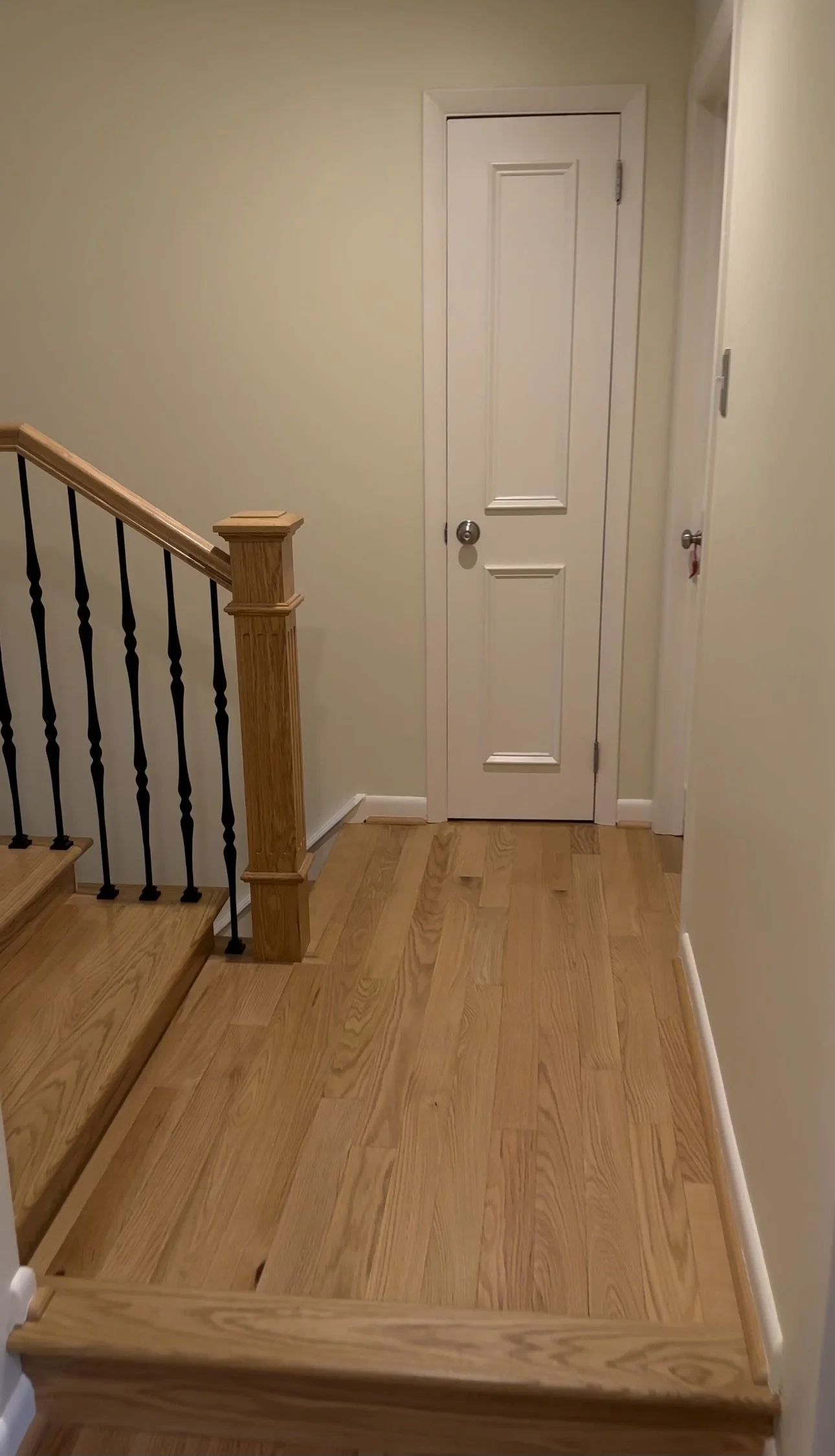 Interior view of a staircase with a wooden handrail and spindles, leading up to a closed white door in a home with light-colored walls and wooden flooring.