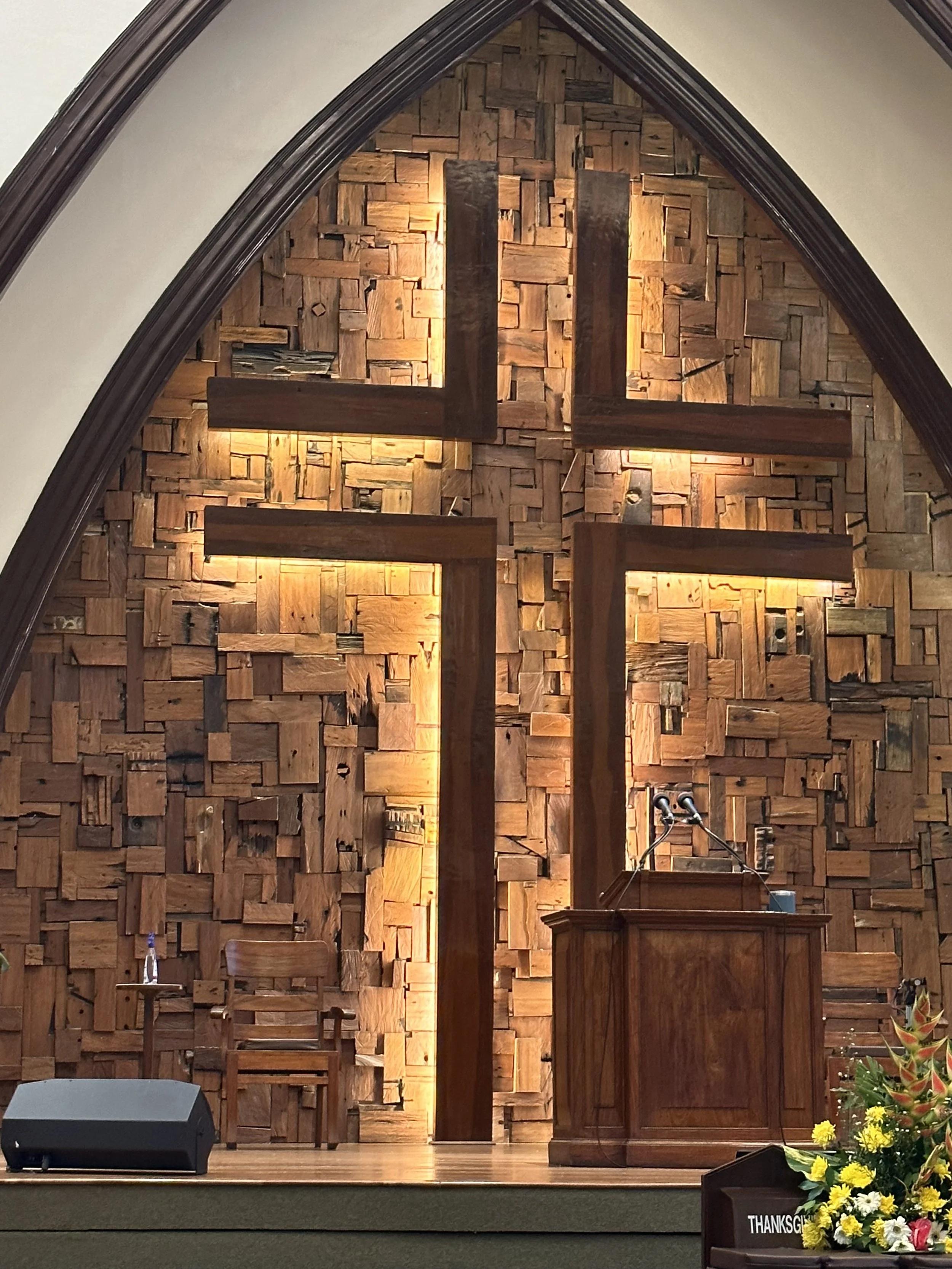 Interior view of a church or chapel with a wooden cross on a wall made of wooden blocks; a podium with a microphone, a chair, and floral arrangements with yellow and red flowers at the front.