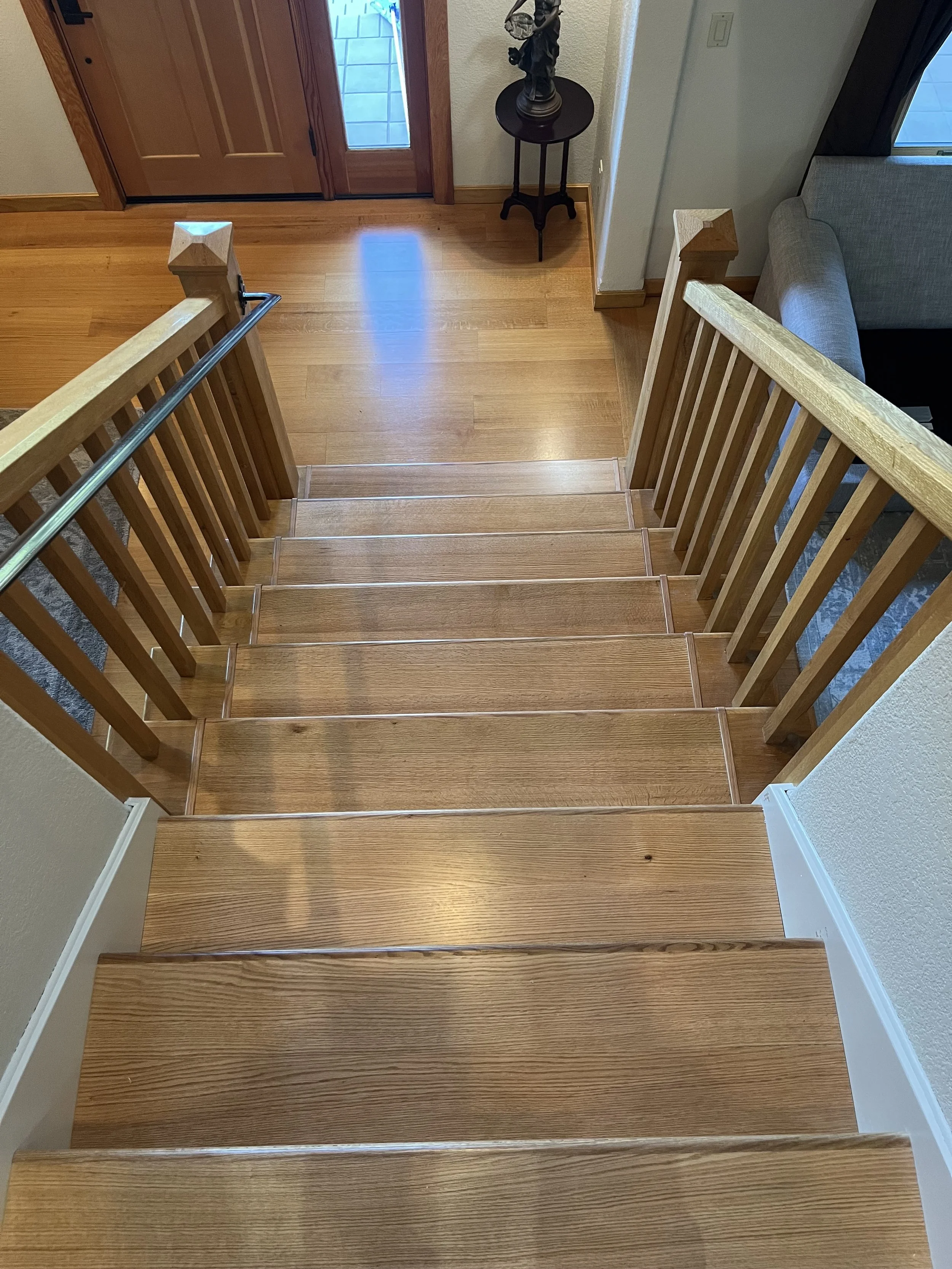 Photo of a wooden staircase seen from the top, with a handrail on the left side. The staircase leads down to a hardwood floor near a wooden front door with a window panel. To the right, part of a beige sofa and a window with a curtain are visible. Th