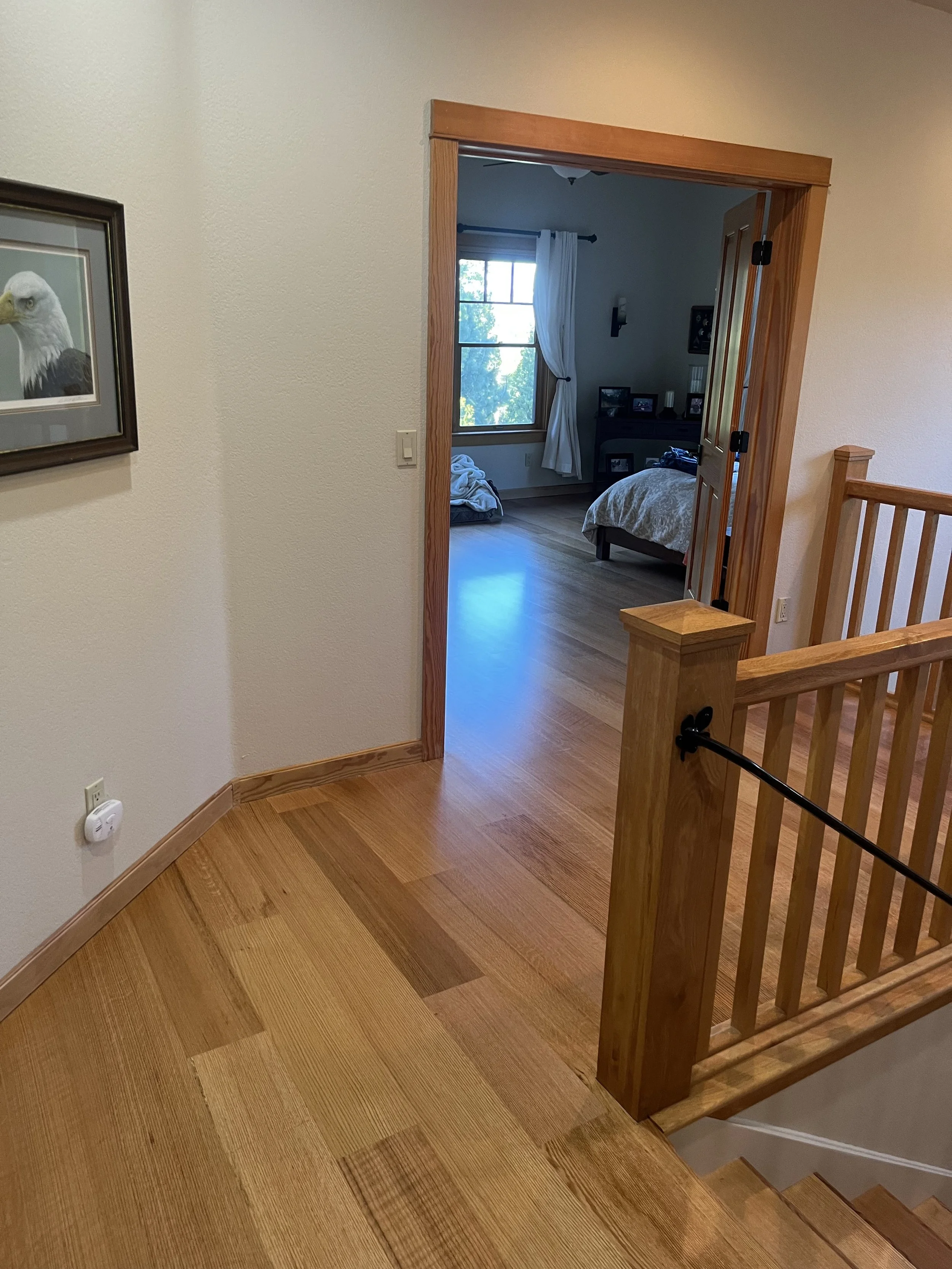 View of a wooden staircase and floor leading to a bedroom with a window, bed, and picture frames. Part of a hallway with a framed eagle picture on the wall.