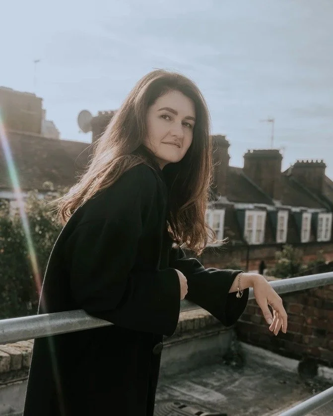 A young woman with long brown hair leaning on a rooftop railing, with city buildings and rooftops in the background, and sunlight creating a lens flare.
