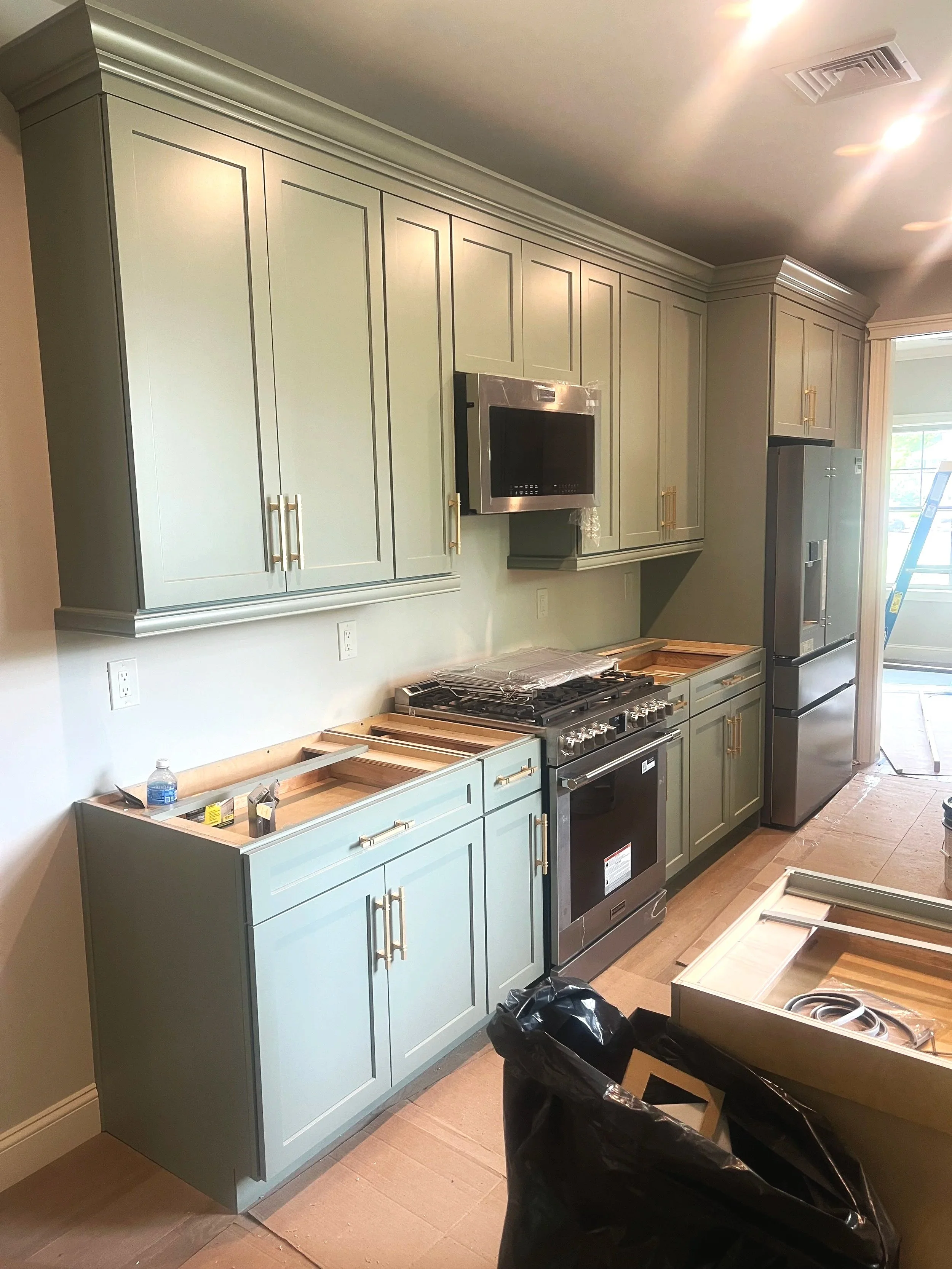 Kitchen with light green cabinets, some without countertops, a stove, and a refrigerator. There is a ladder outside a window and some construction materials and trash bags in the kitchen.