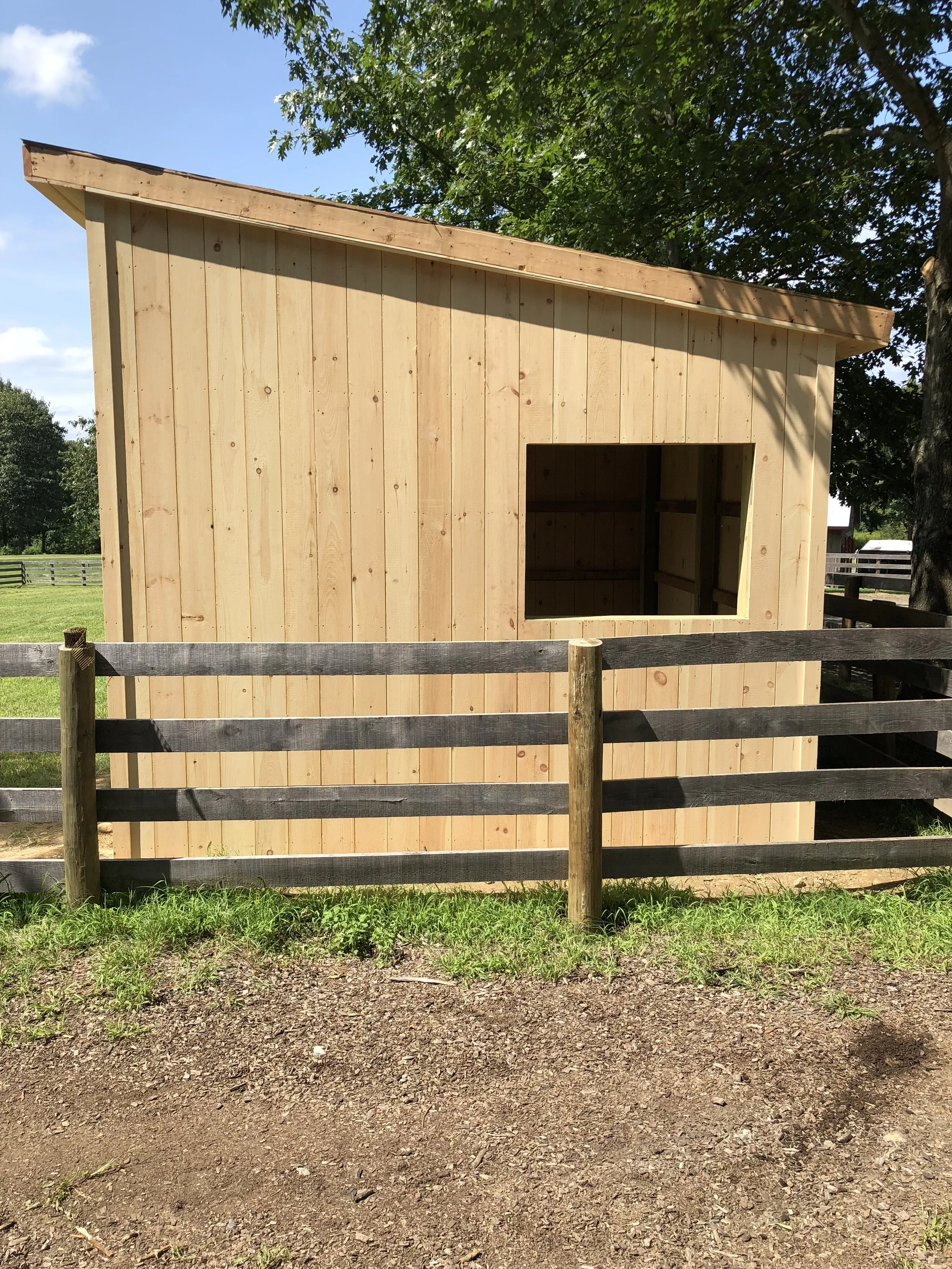 A wooden shed under construction with an open window and a sloped roof, surrounded by a wooden fence and green grass, with trees and blue sky in the background.