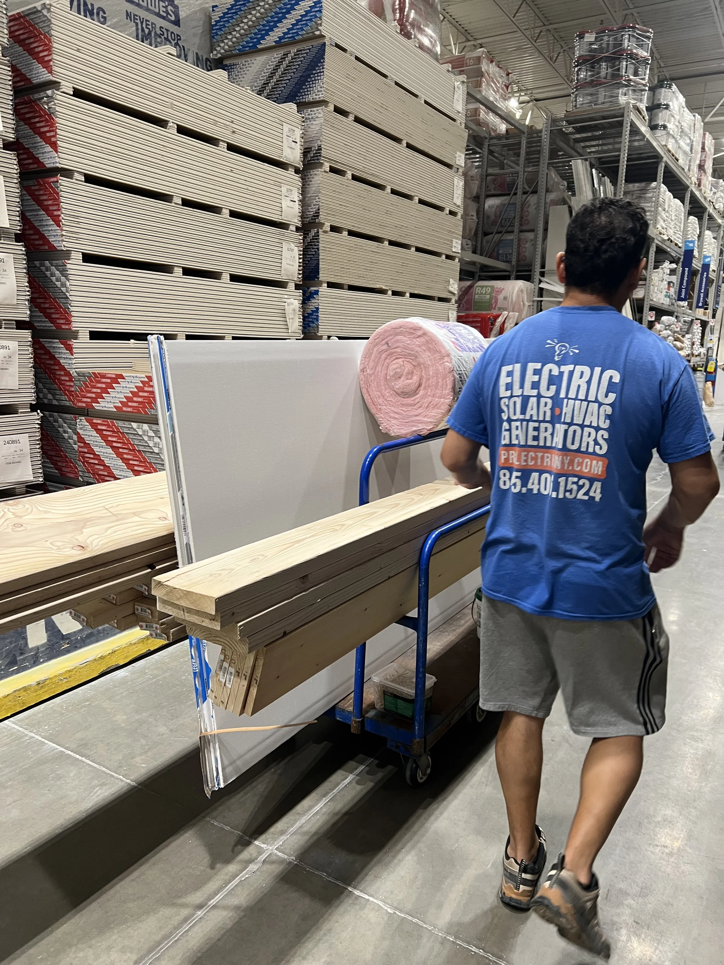 A person wearing a blue t-shirt and shorts is shopping in a hardware store aisle. The aisle contains various types of wood, drywall, insulation, and construction materials. The person is seen from behind, pushing a blue cart.