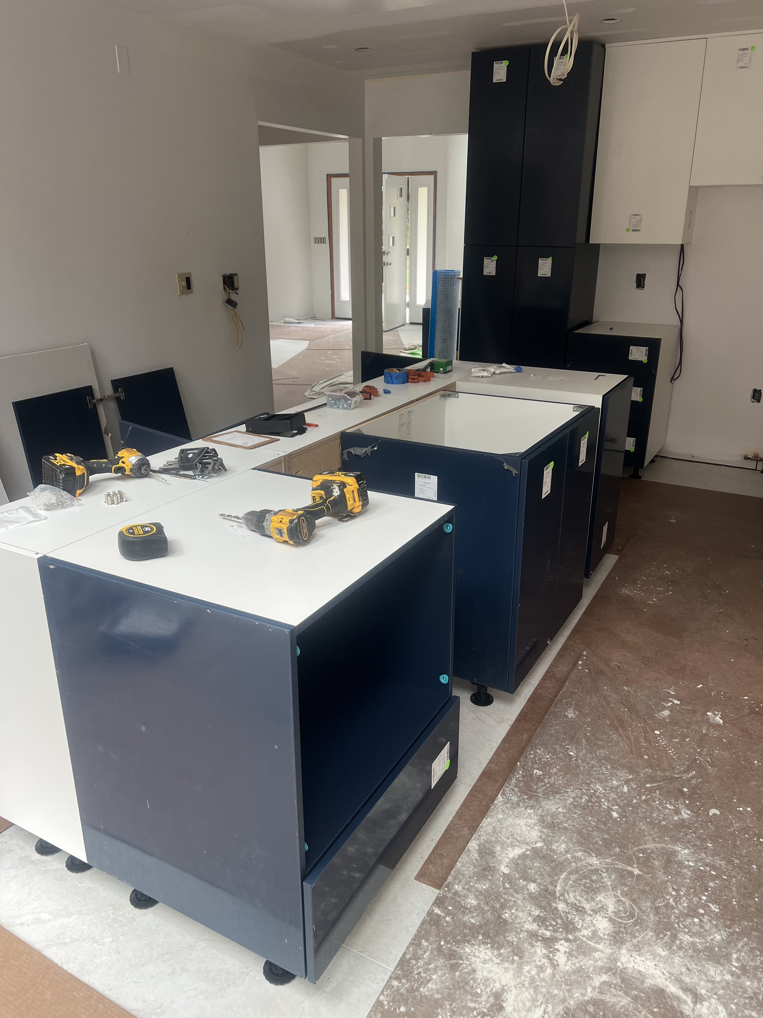 Interior of a kitchen under construction with cabinets and tools on the counter. The floor shows white and brown tiles, with some construction dust.
