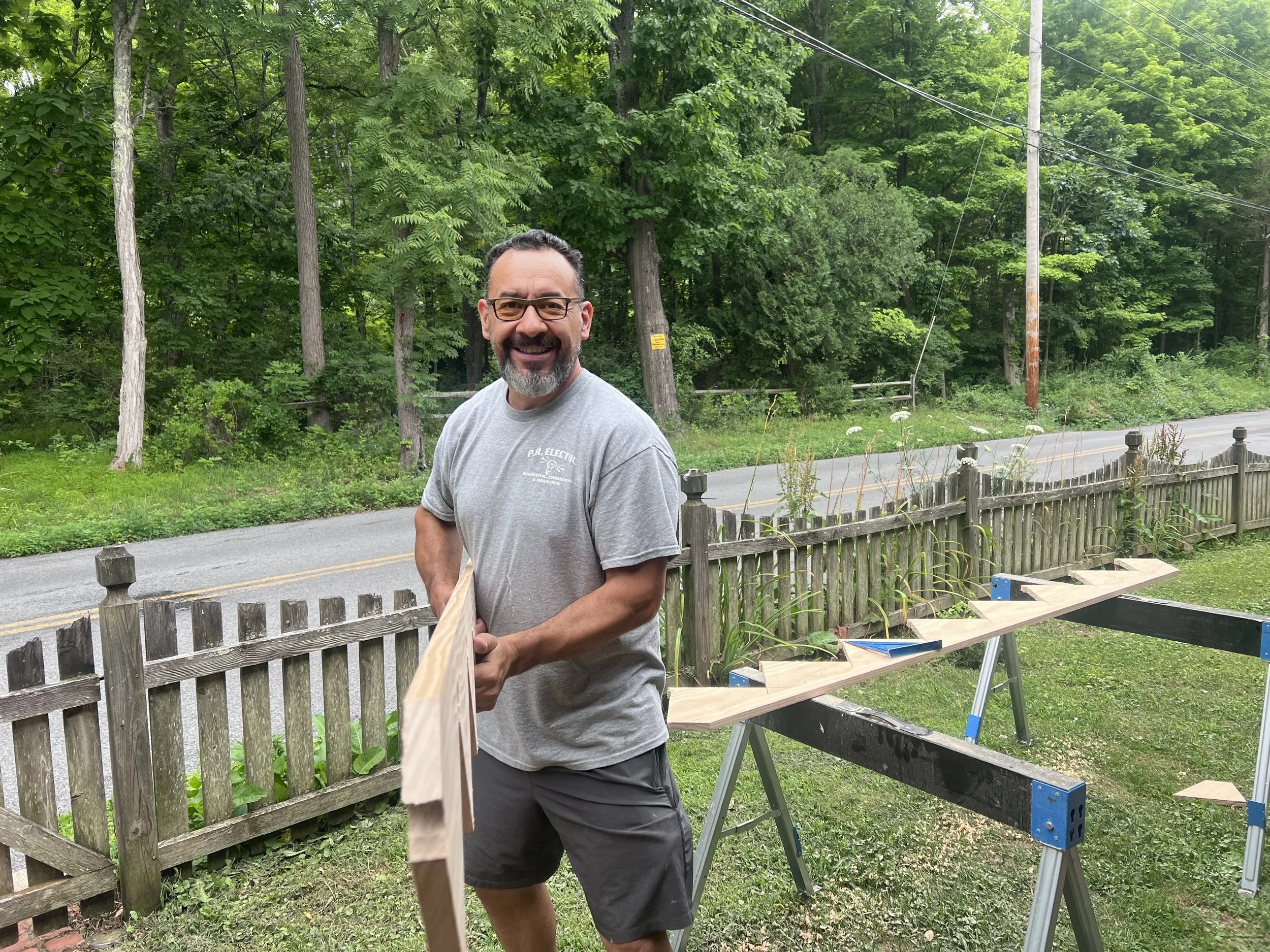 A man with glasses and a beard smiling outdoors, working on a woodworking project with a workbench and wooden planks, in front of a wooded area with a road and utility poles.