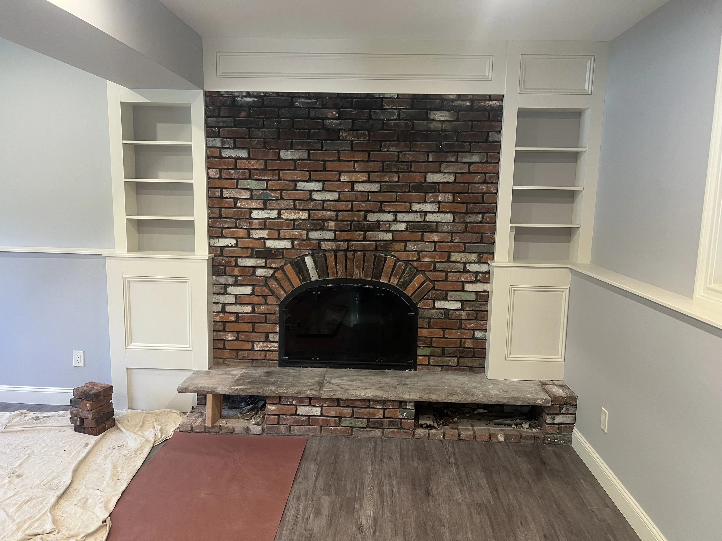 Interior of a room with a brick fireplace under construction, surrounded by built-in white shelves, with construction materials like bricks and fabric on the floor.