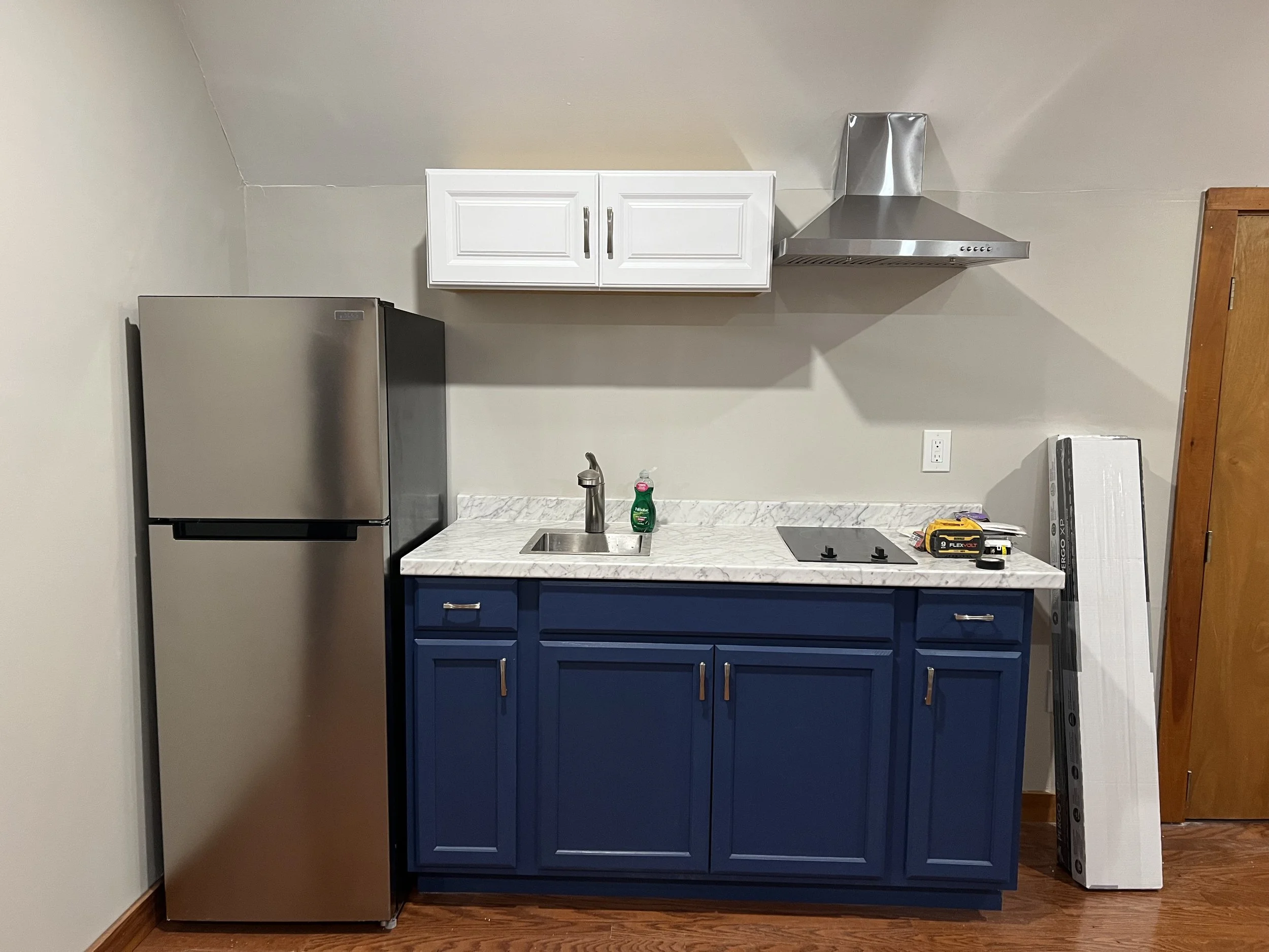 Kitchen with silver refrigerator, white upper cabinets, stainless steel range hood, dark blue lower cabinets, marble countertop, sink, dish soap, cordless drill, box, fan, and a towel on a wooden floor.