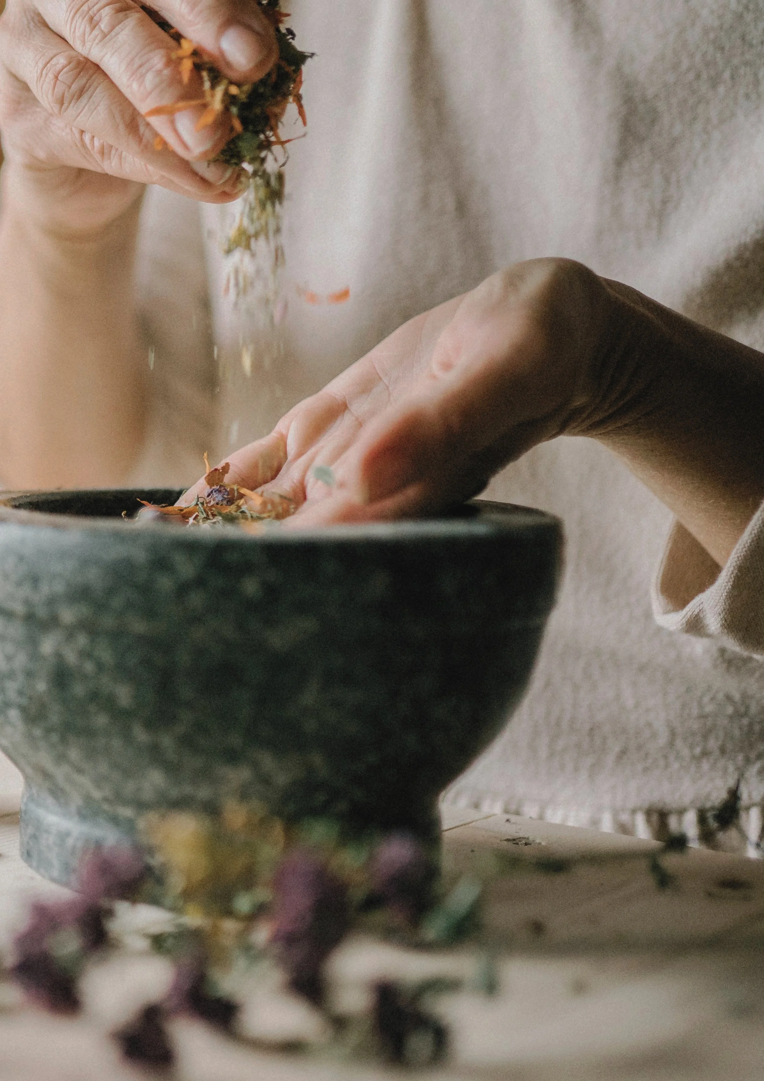 Close-up of a person's hand holding dried herbs or flowers over a large dark stone bowl, with blurred flowers in the foreground.