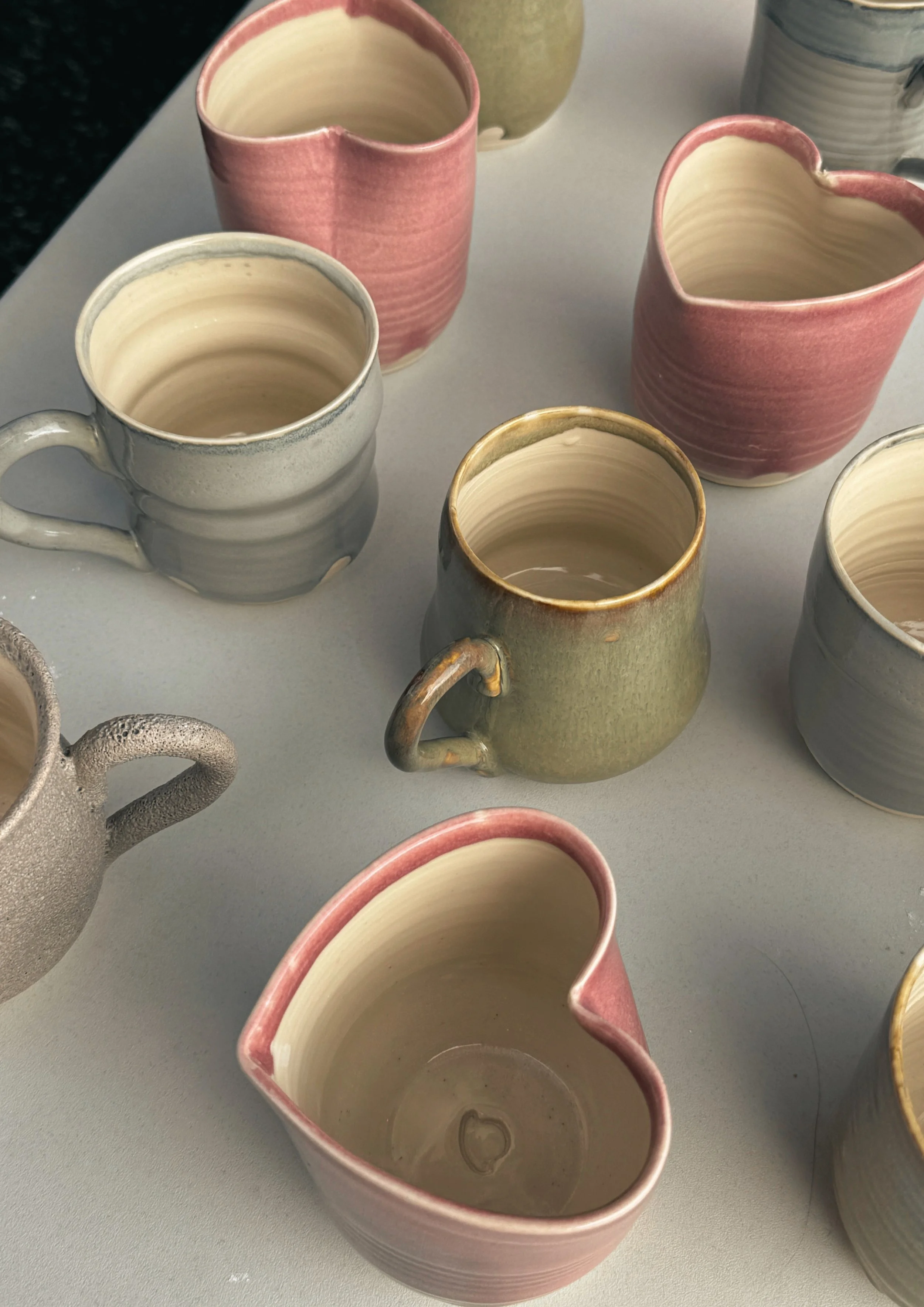 Multiple ceramic mugs and pitchers with glazed and unglazed finishes in pastel colors on a white surface.