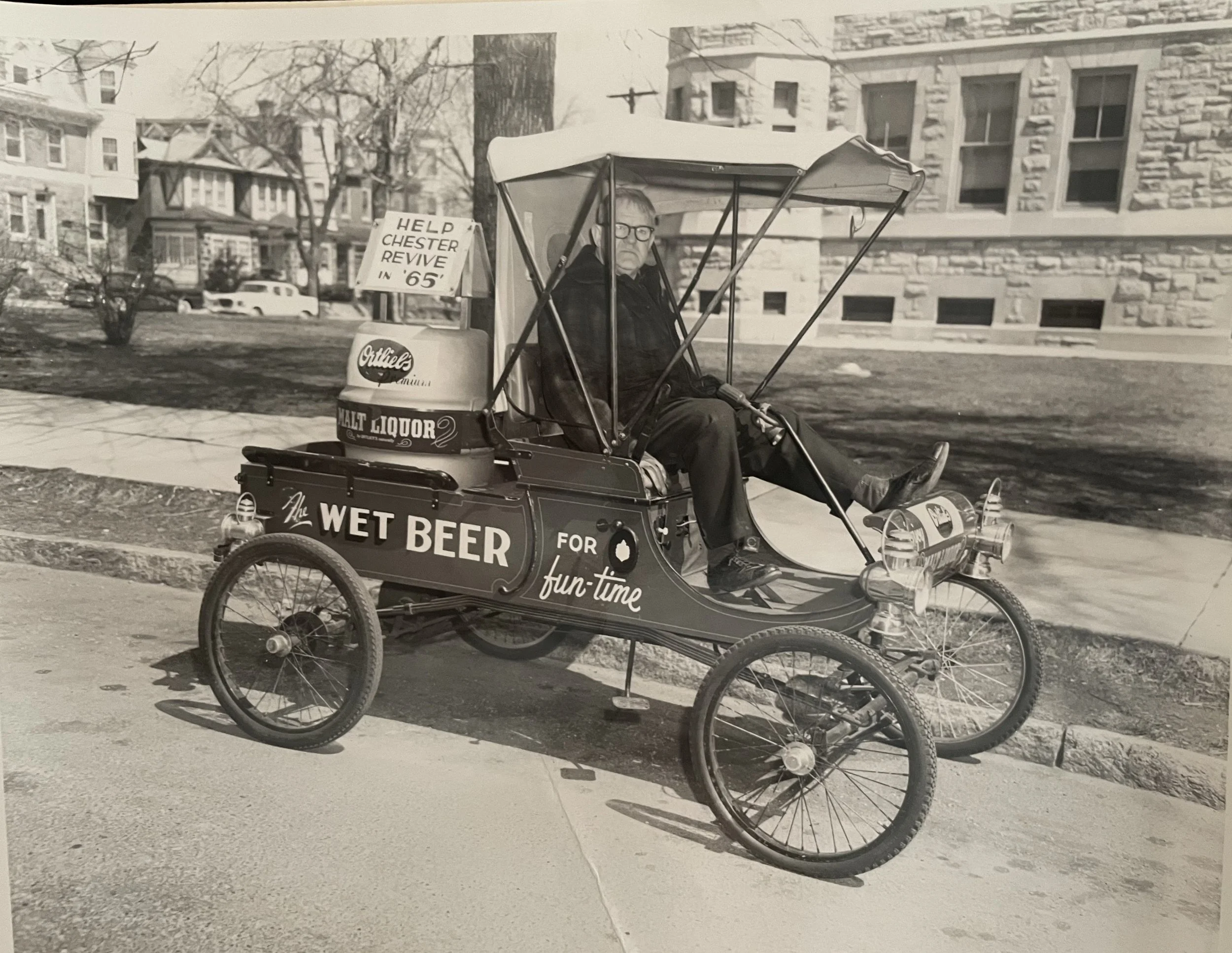 A photo from 1965 showing a man drive through Chester in an Ortlieb's beer truck probably dating from the 1910's (or even earlier). Notice the lack of steering wheel and the little horn attached as well. Handy little truck to have with you for the bi
