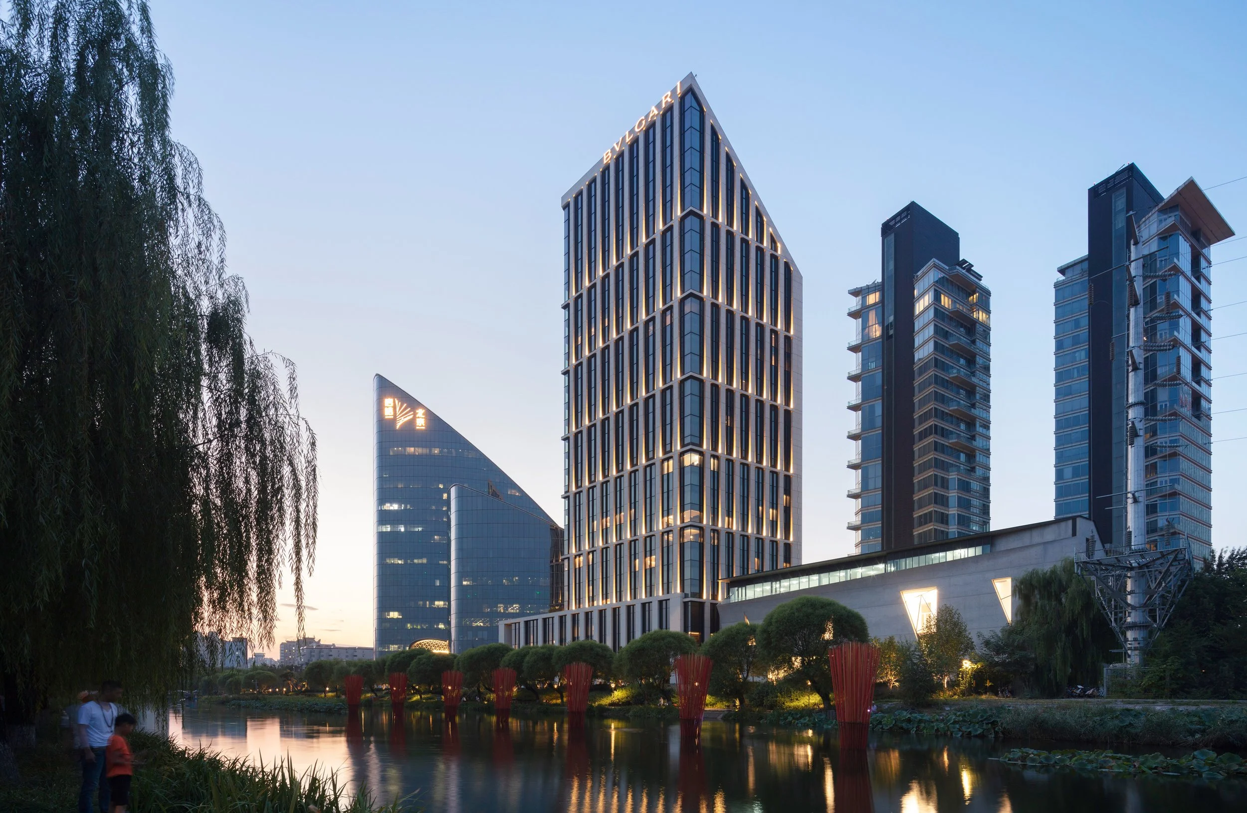 City skyline at dusk featuring tall modern buildings with illuminated windows overlooking a park with a pond, trees, and people walking nearby.