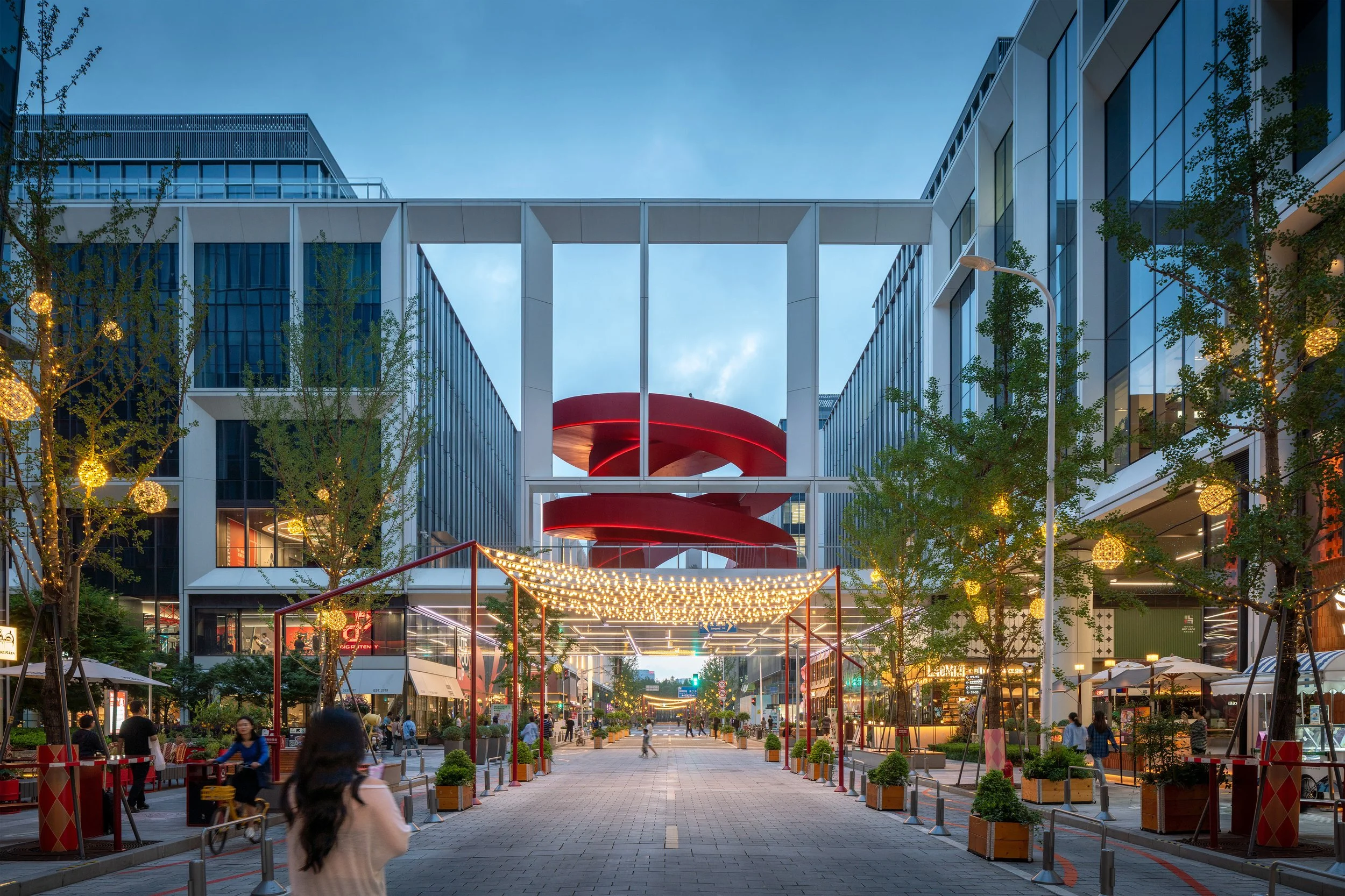People walking in an outdoor shopping area with modern glass buildings, decorated with string lights and trees, during evening.