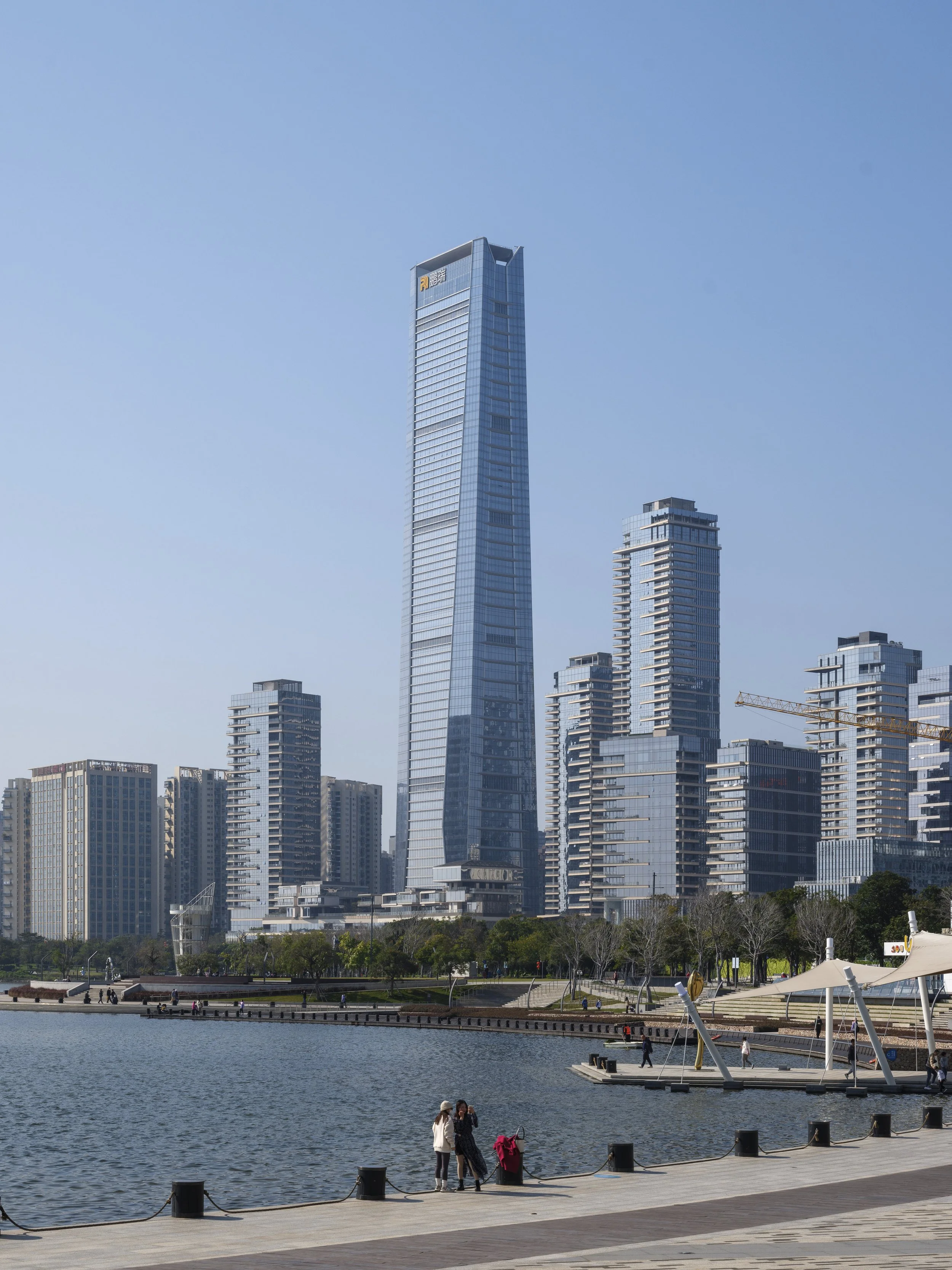 A cityscape with tall modern skyscrapers seen from a waterfront park with a walkway and benches, and people walking and sitting beside the water.