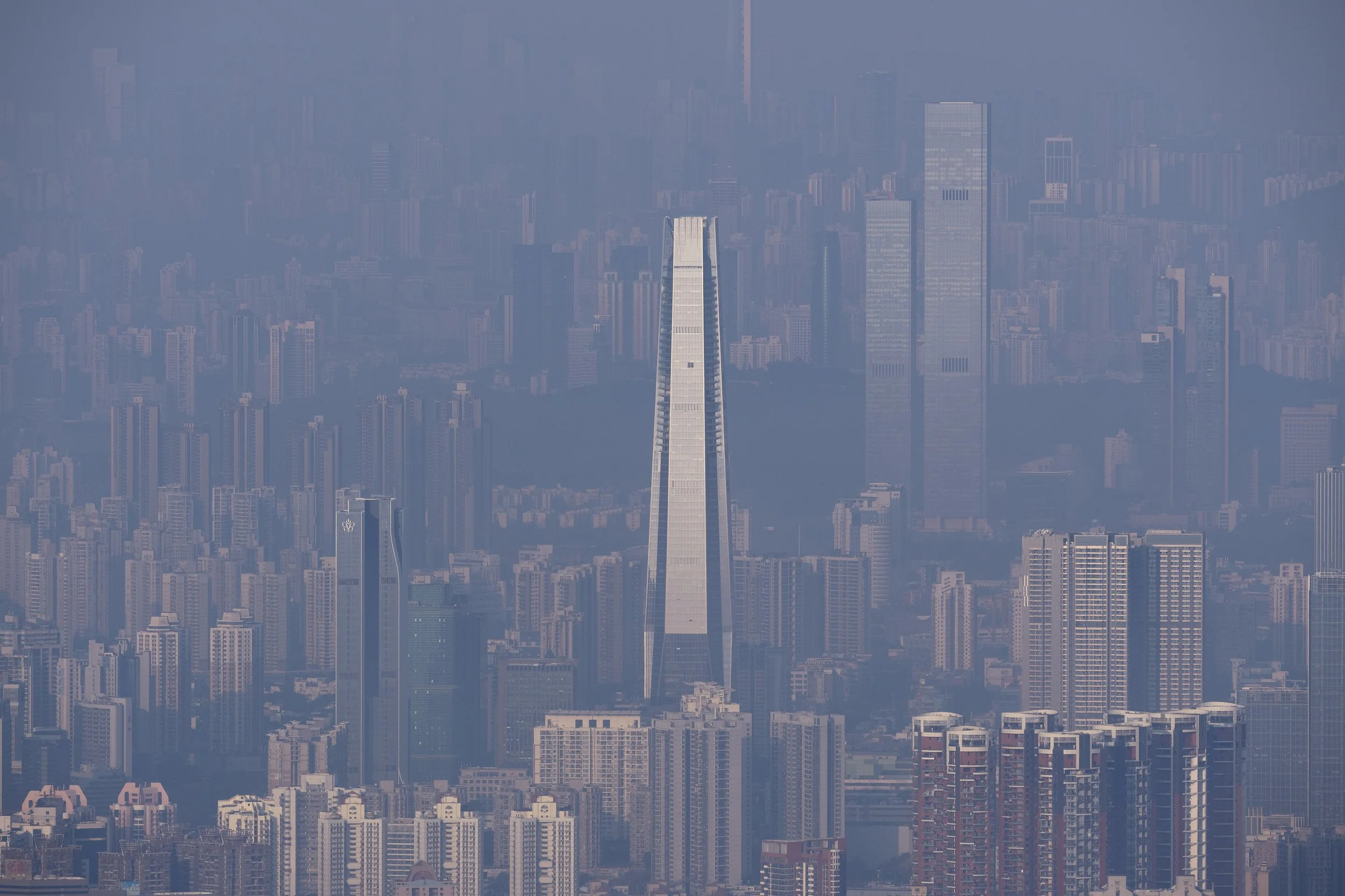Aerial view of a city with many tall skyscrapers and buildings, featuring the tallest pointed building in the center, surrounded by a hazy sky.