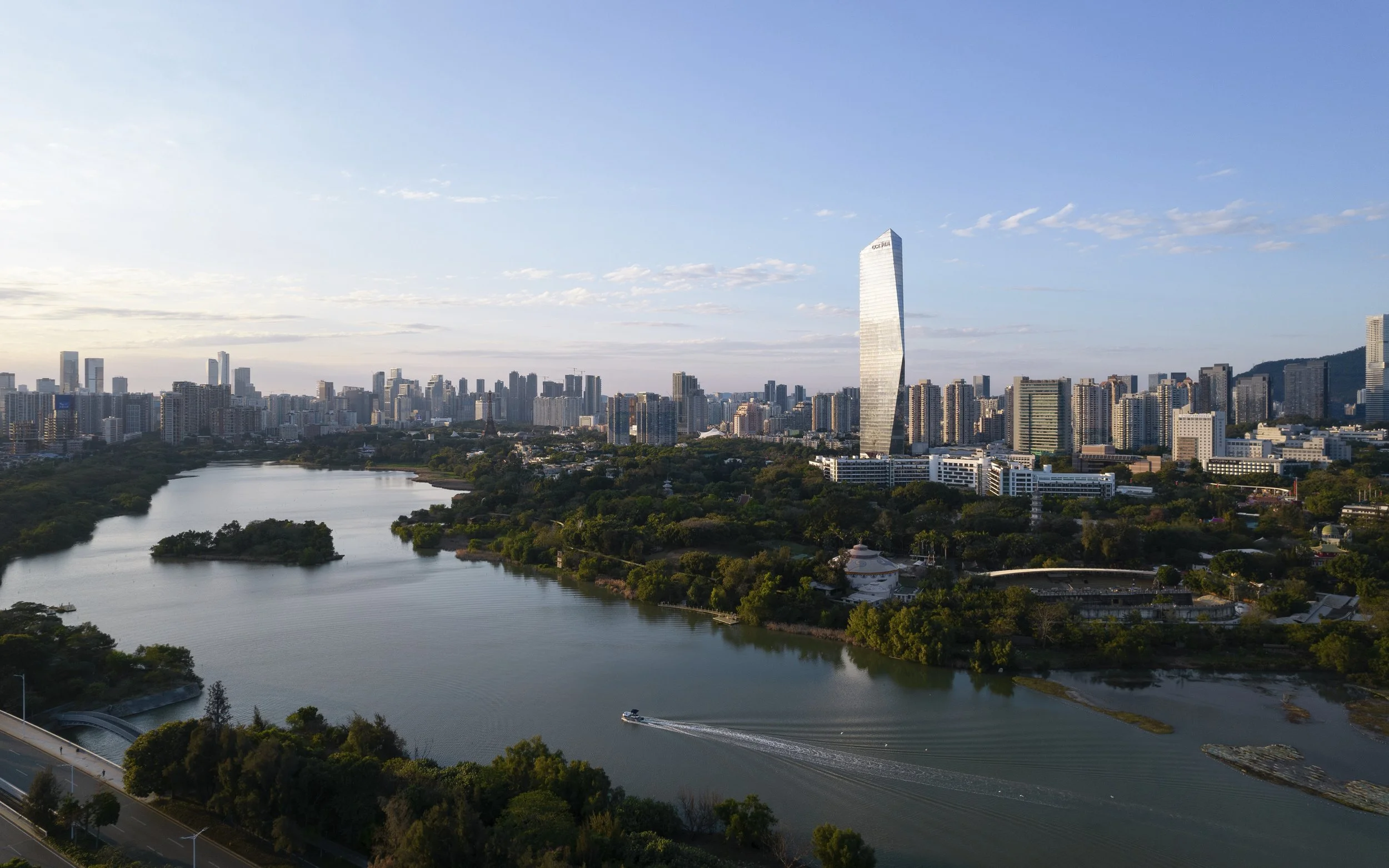A city skyline featuring a tall, modern skyscraper with a slight twist in its design, near a large body of water with greenery and trees around it, and a boat leaving a wake in the water, under a partly cloudy blue sky.