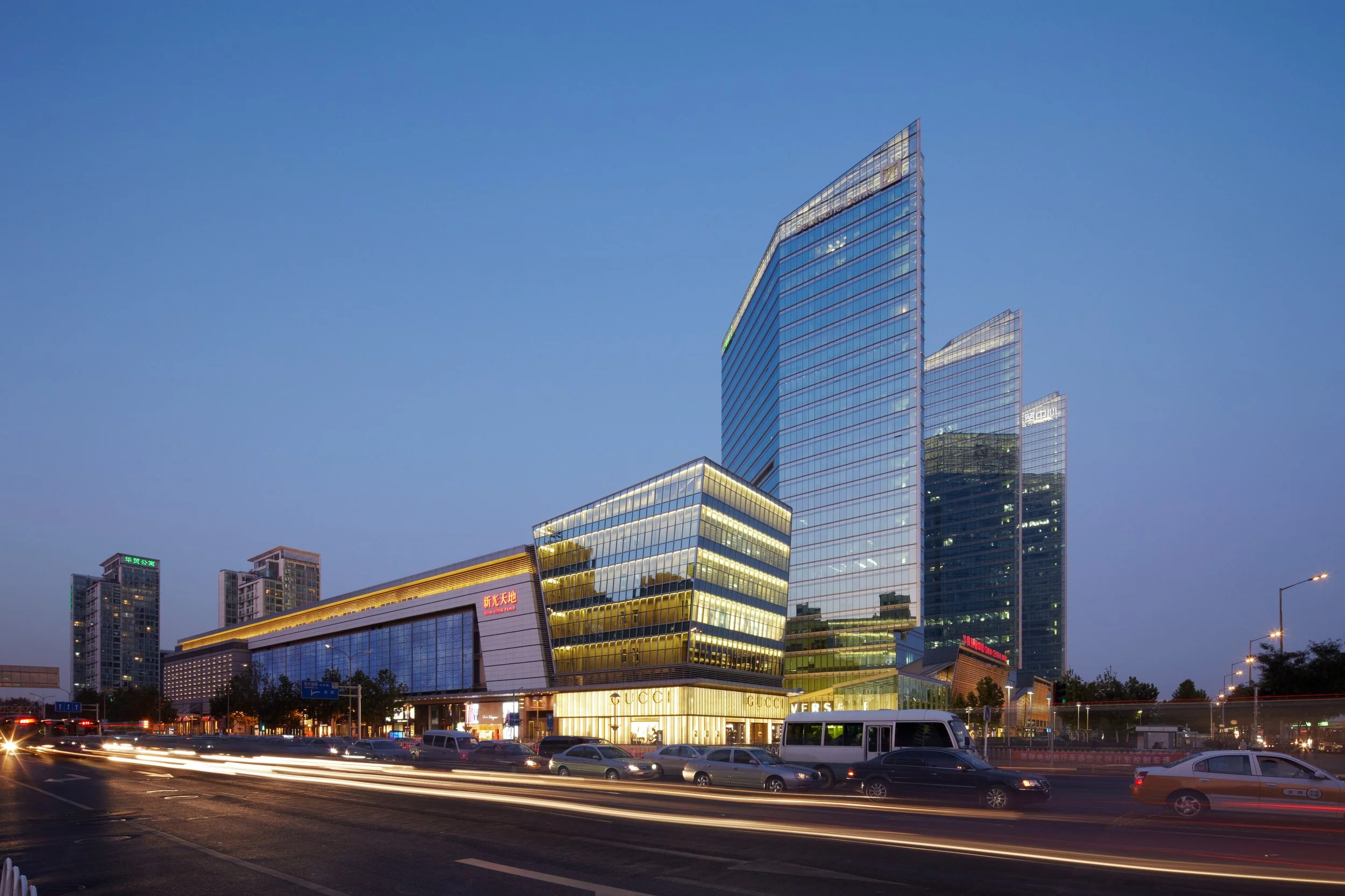Modern glass skyscrapers and commercial buildings in an urban cityscape at dusk, with cars passing by on the street and light trails from moving vehicles.