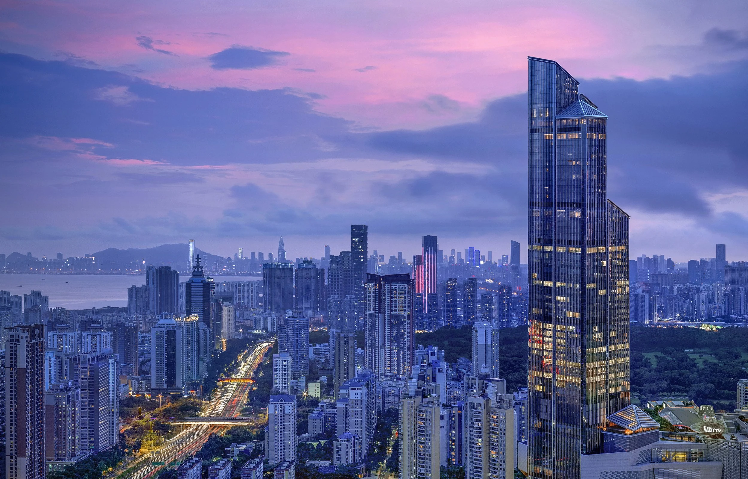 Nighttime city skyline with high-rise buildings, some with illuminated windows, under a cloudy sky with pink and purple hues.