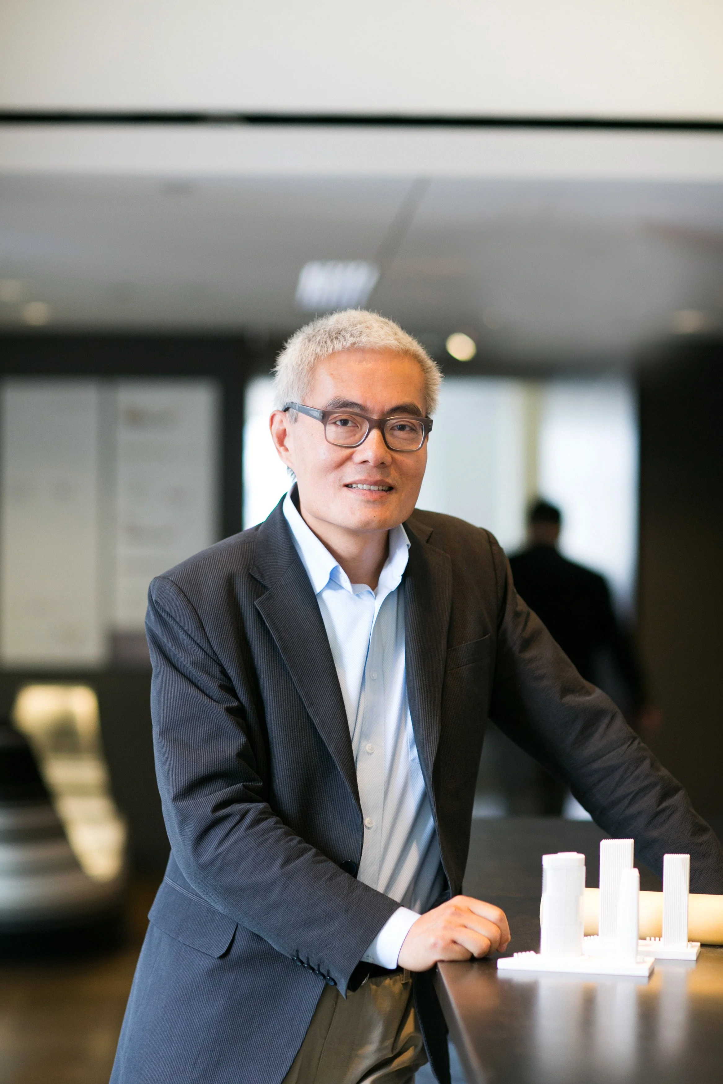 A man with glasses, wearing a suit, leaning on a table with white architectural models in an office or conference room.