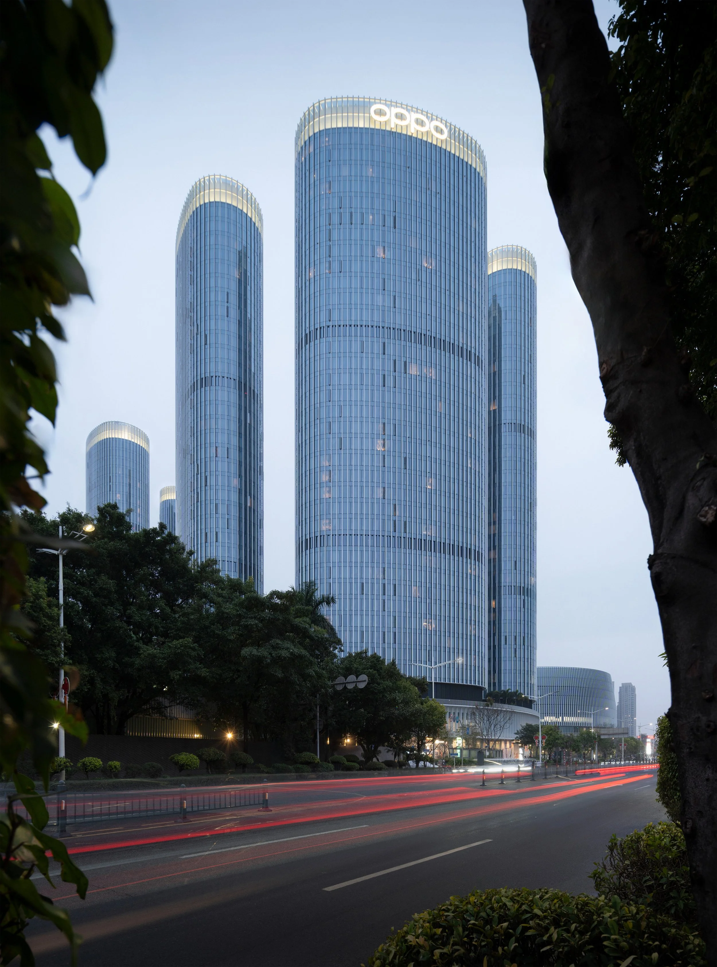 Modern cityscape with tall, glass-covered skyscrapers and the 'Oppo' logo at the top of the central building, framed by trees and streetlights at dusk.