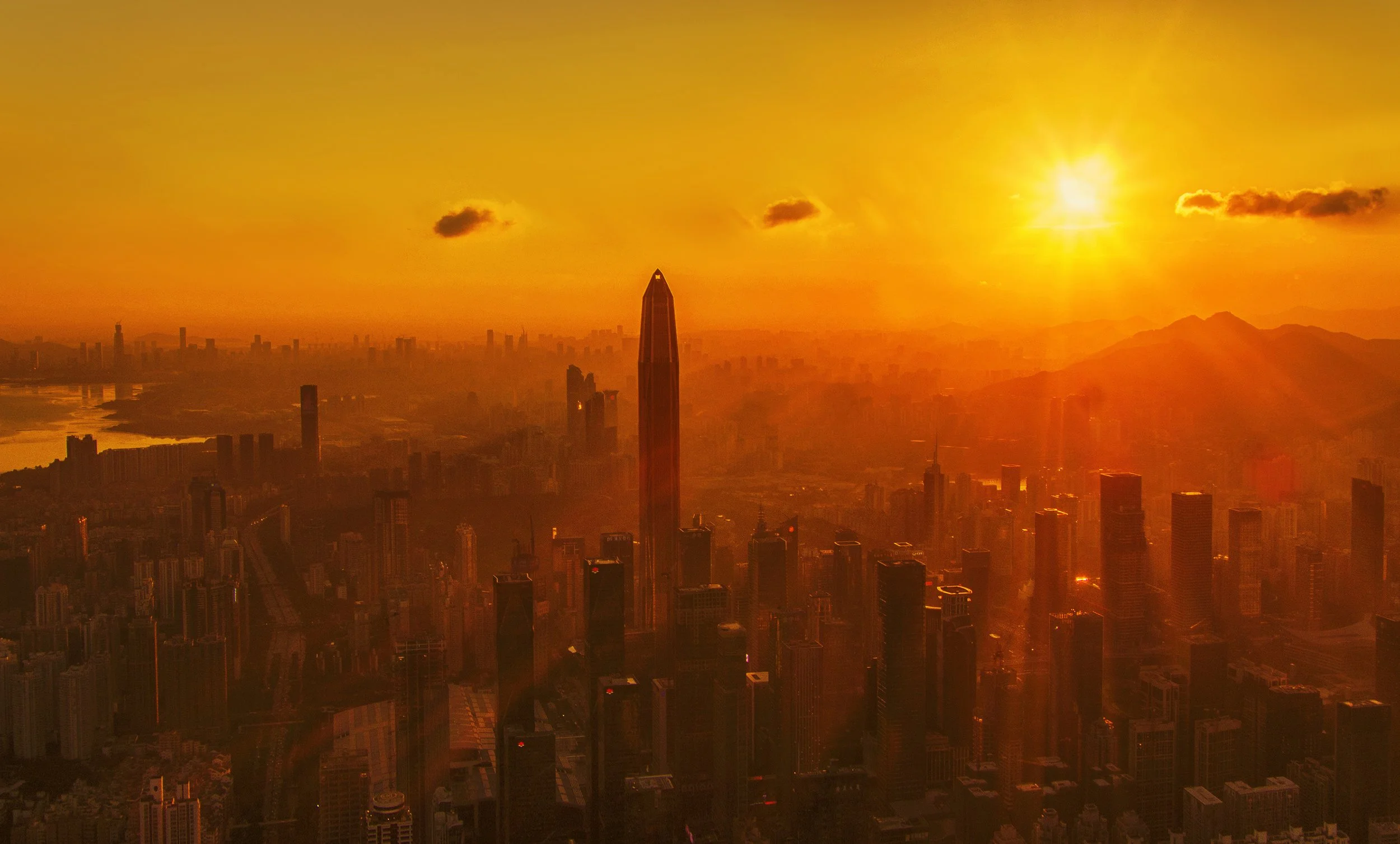 City skyline at sunset with tall buildings and mountains in the background, orange sky, and the sun near the horizon.