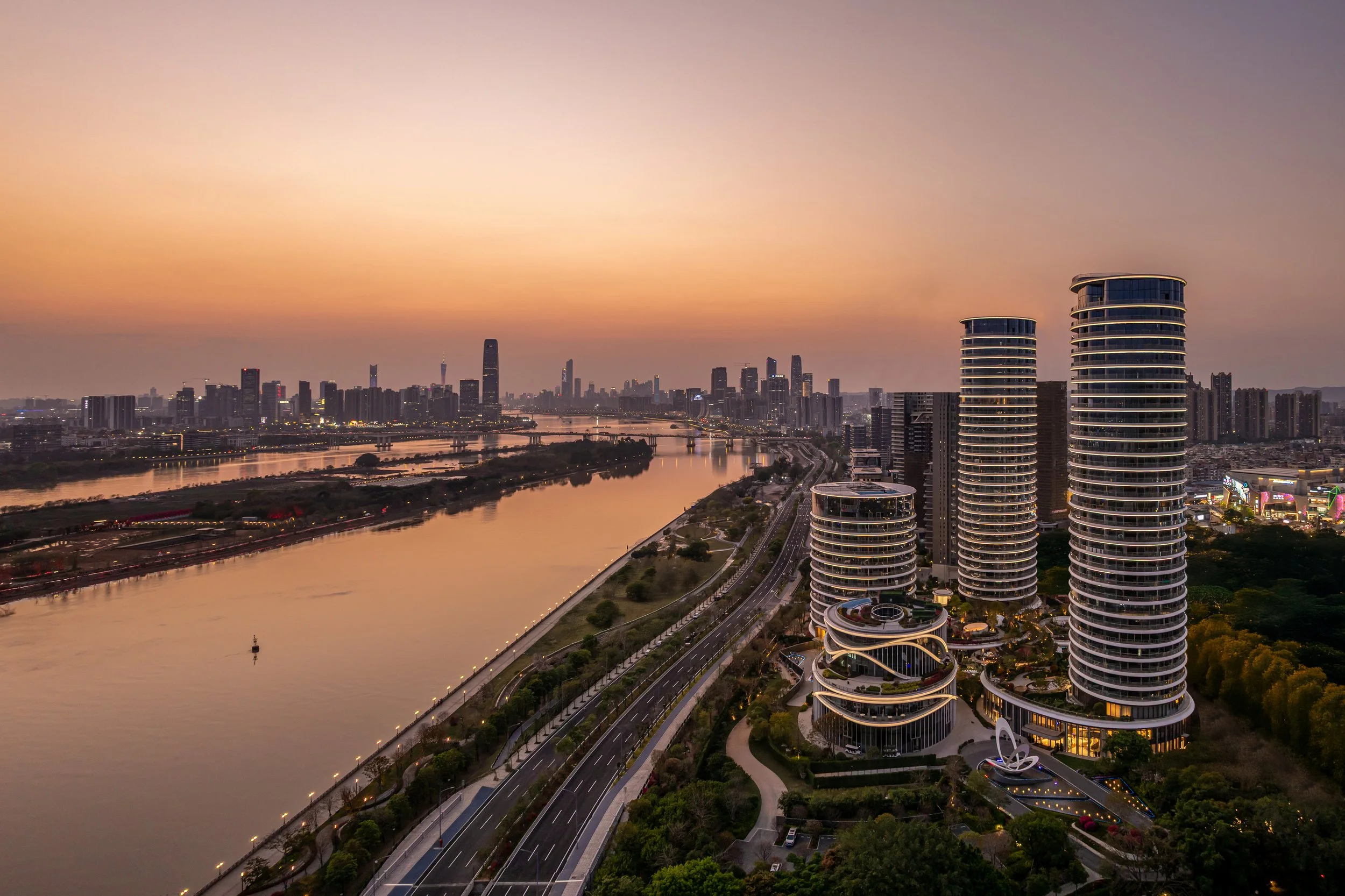 A city skyline at sunset with tall buildings, a river reflecting city lights, and a distinctive curved residential building in the foreground.