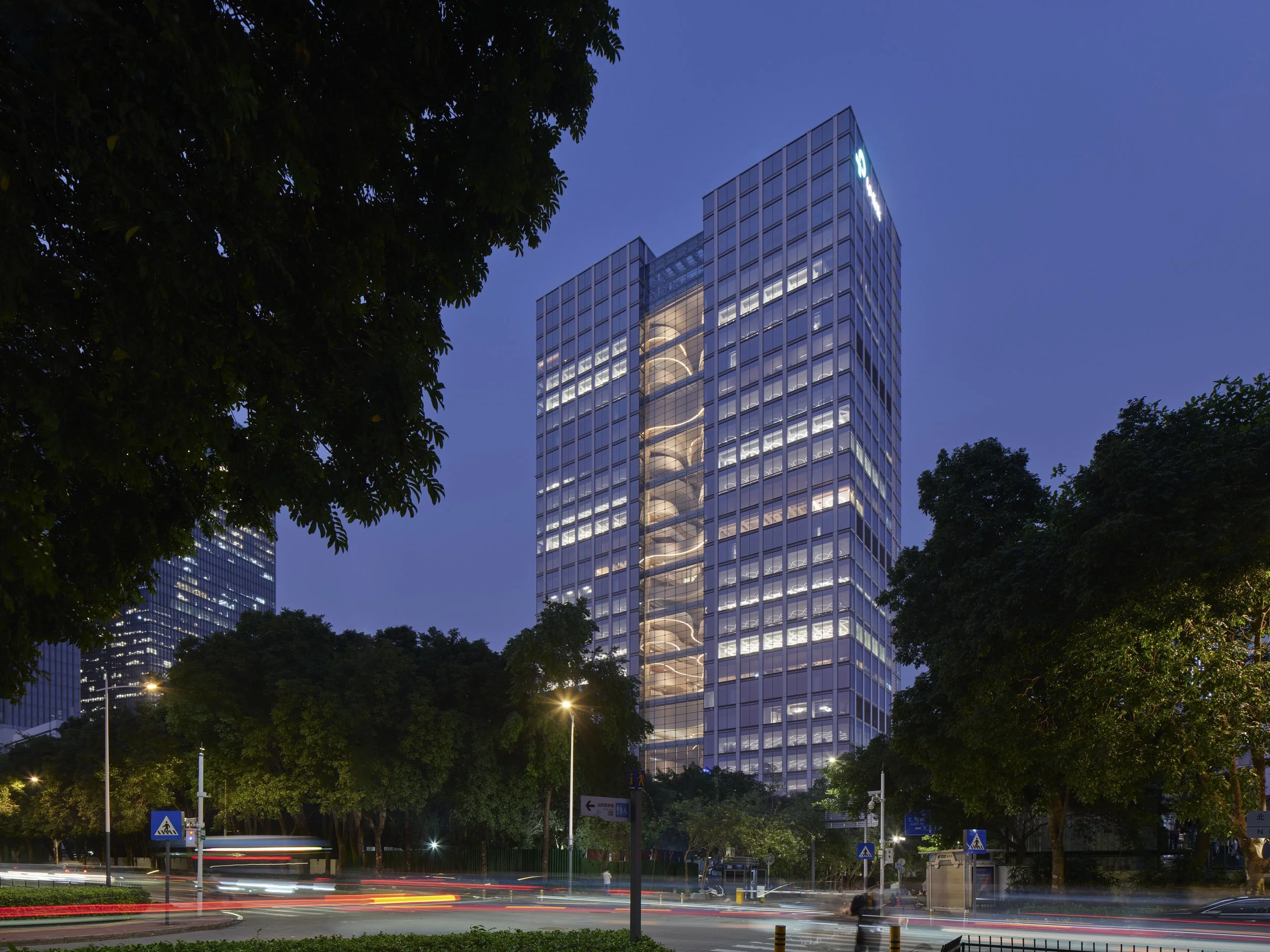 Night view of tall glass office building with illuminated windows, surrounded by trees and streetlights. Light trails from moving vehicles are visible on the street in front of the building.