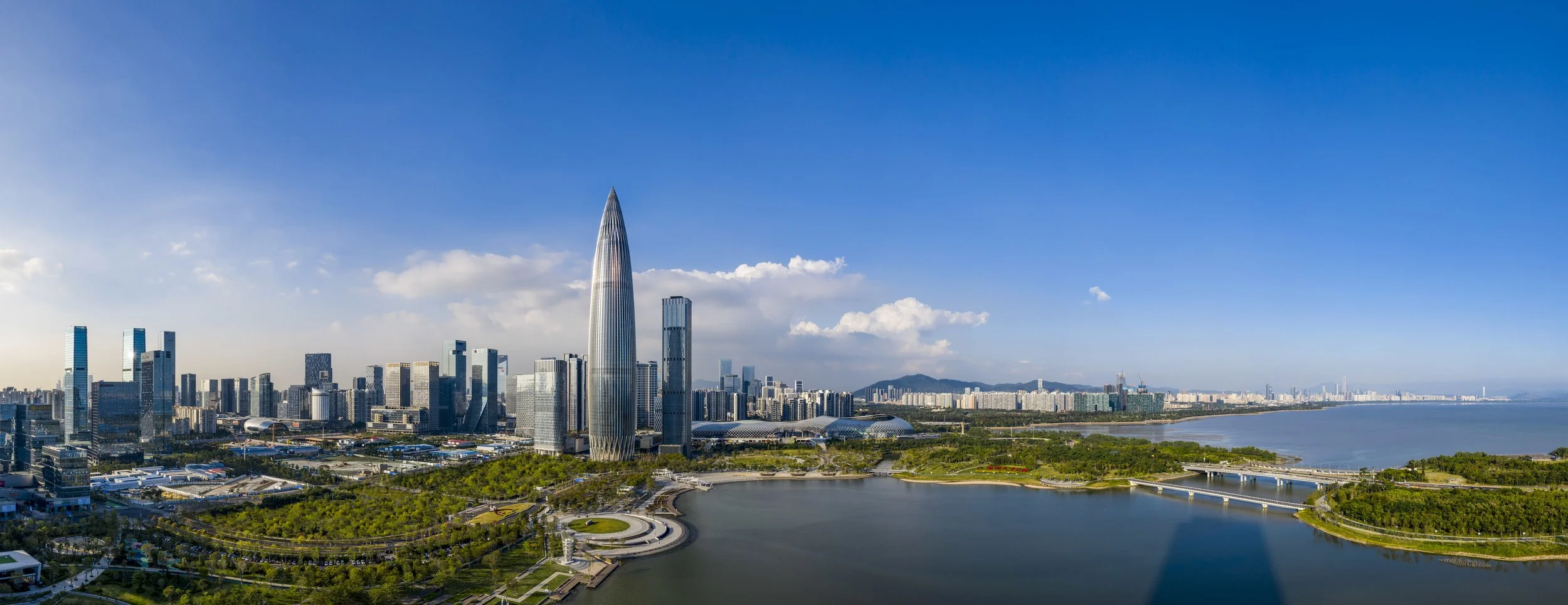 A city skyline with modern skyscrapers, including a tall, pointed building, next to a body of water on a clear, sunny day.
