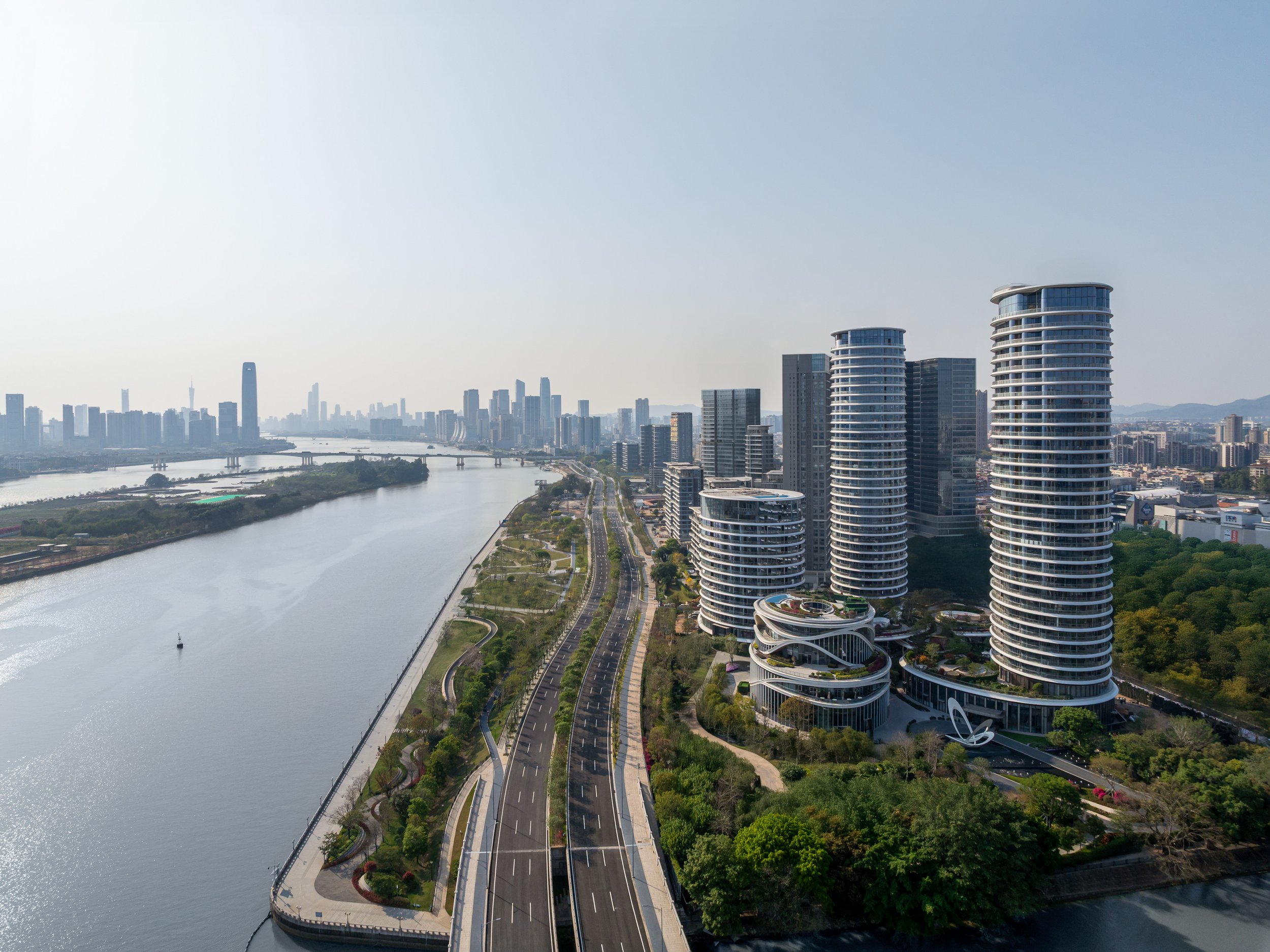 Aerial view of modern skyscrapers along a river with a bridge, featuring two tall, uniquely shaped residential buildings with curved glass facades, and a distant city skyline under a clear sky.
