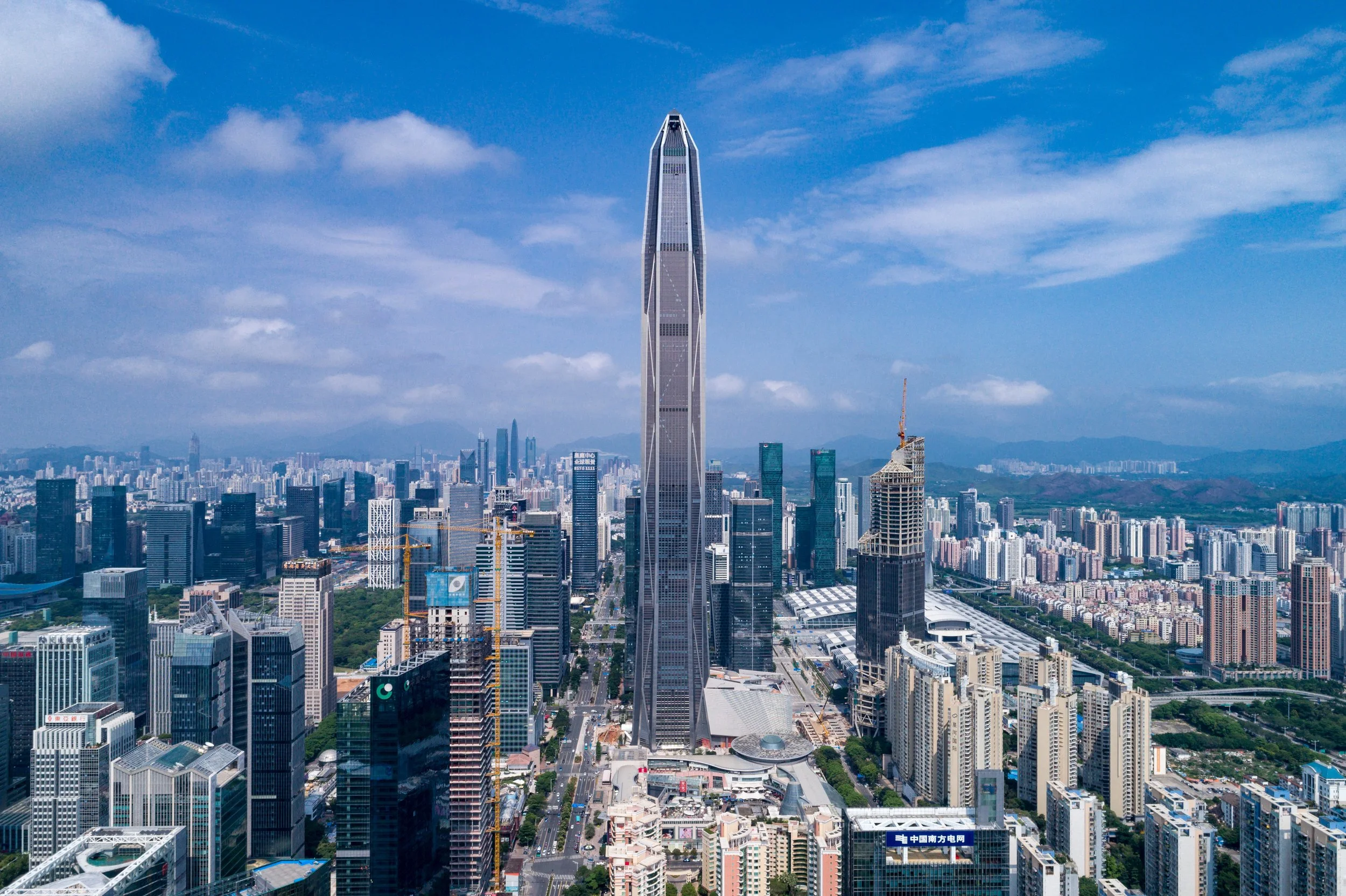 Skyline of a modern city with a tall, distinctive skyscraper in the center, surrounded by other high-rise buildings and construction cranes, under a blue sky with scattered clouds.