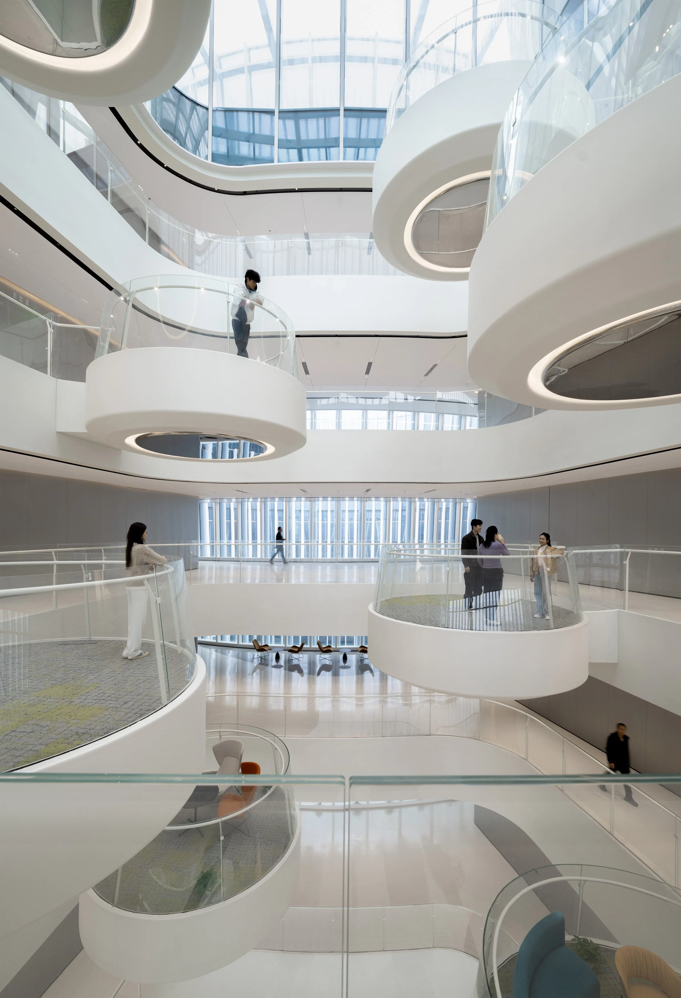 Interior view of a modern, multi-level building with curved white balconies and glass railings. Several people are walking and standing, including a person on the upper balcony looking down. There is natural light coming through a large glass ceiling and windows, with a spacious, airy atmosphere.