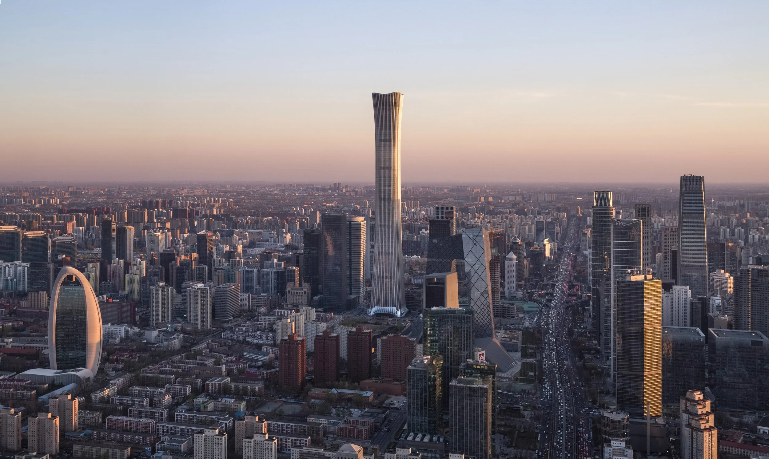 Aerial view of a city skyline during sunset, featuring tall skyscrapers including a curving tower, a large white arch structure, and numerous other high-rise buildings.