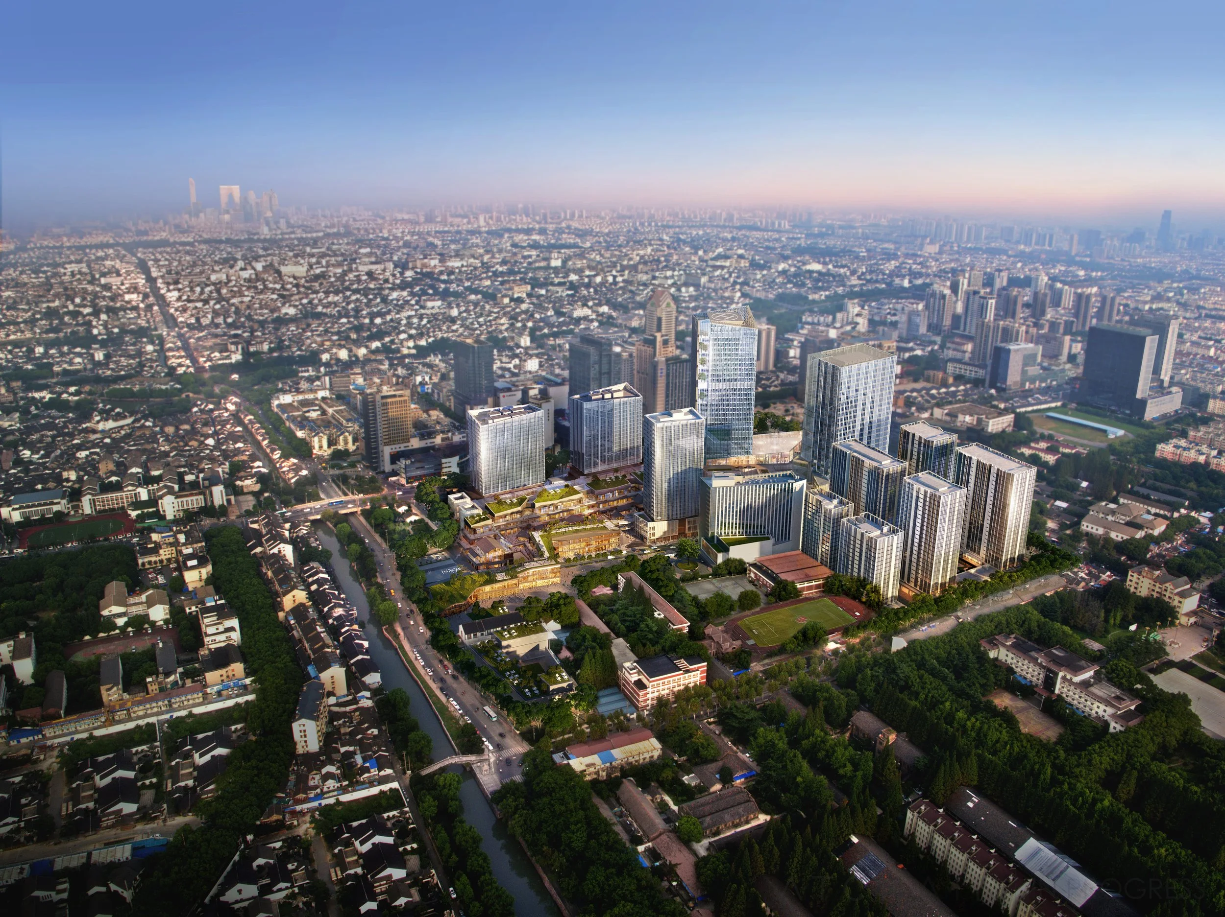 Aerial view of a cityscape at sunset showing many high-rise buildings, dense residential areas, a river, and a sprawling urban landscape with a hazy sky.
