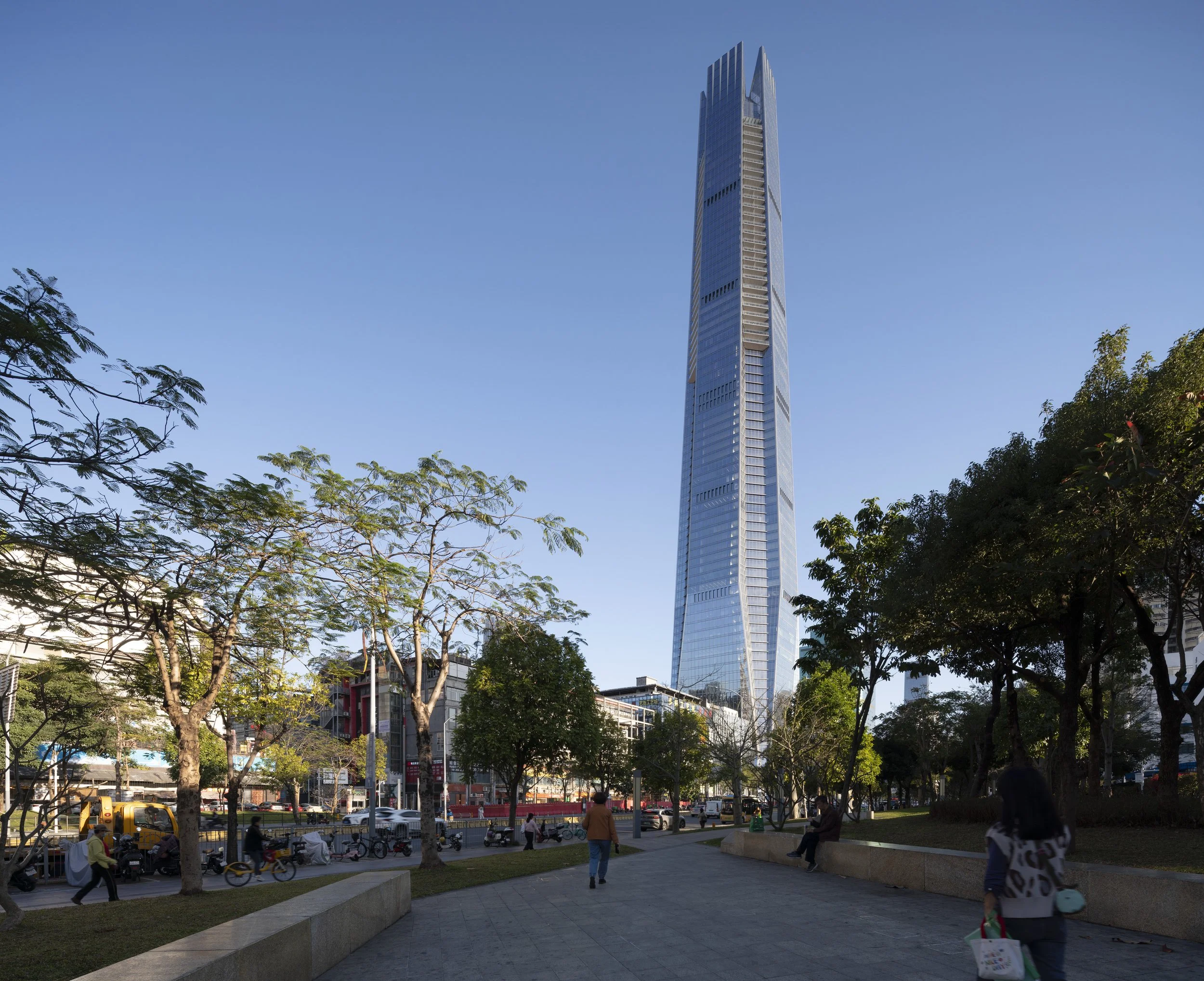Tall modern skyscraper under clear blue sky, seen from park with trees, people, and bicycles