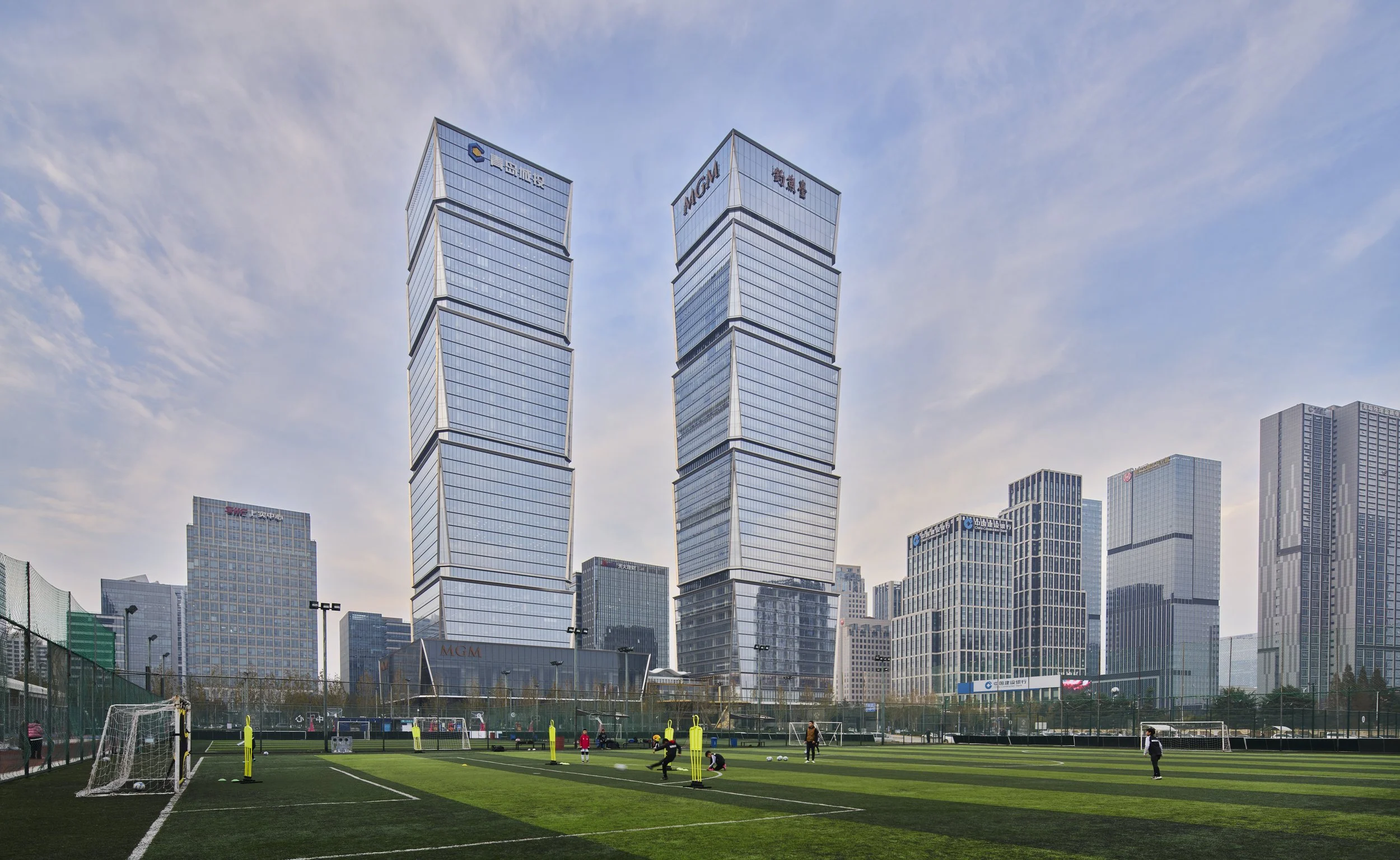 Urban cityscape with modern skyscrapers and a sports practice field in the foreground, where children are playing soccer and participating in athletic activities.
